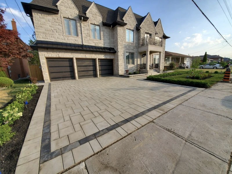 A modern stone house with a large paved driveway, three garage doors, grassy area, and flowering shrubs under a partly cloudy sky.