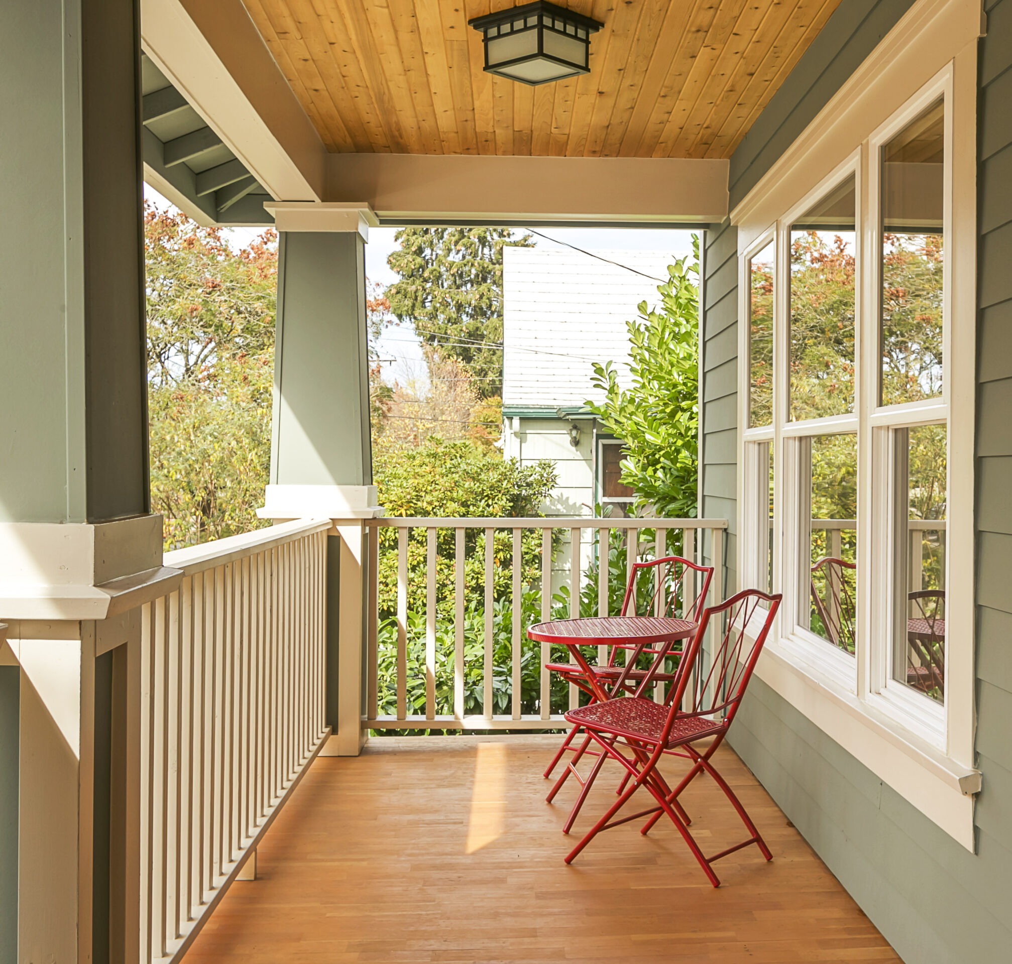 A cozy porch with green siding features red chairs and a small table, surrounded by lush greenery under a wooden ceiling.