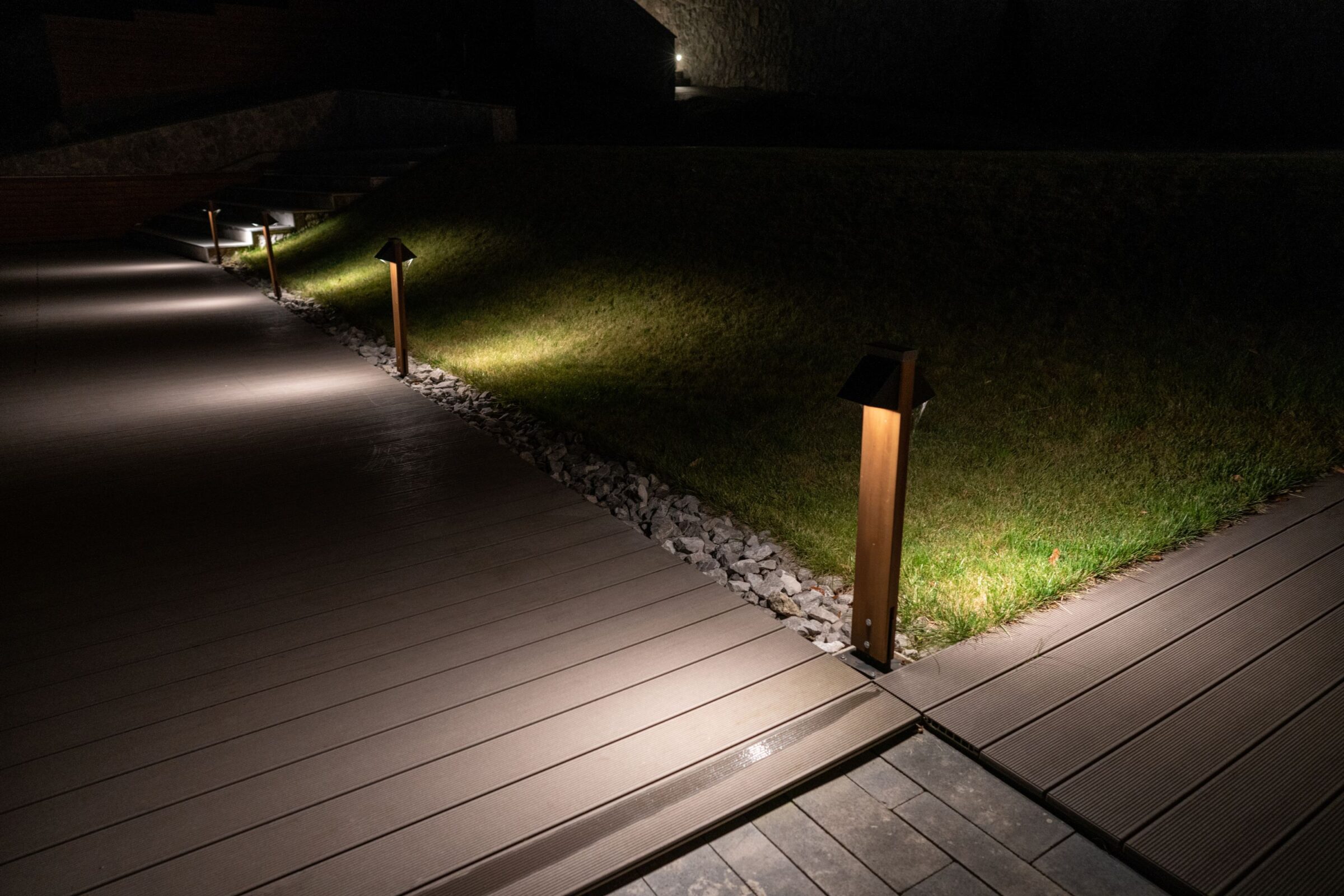 Pathway with illuminated bollard lights, bordered by grass and gravel. Nighttime setting with modern landscaping elements visible along the walkway.