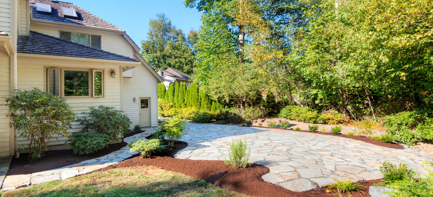 A residential house with beige siding, surrounded by a landscaped garden, stone pathway, trees, and shrubs under bright sunlight.