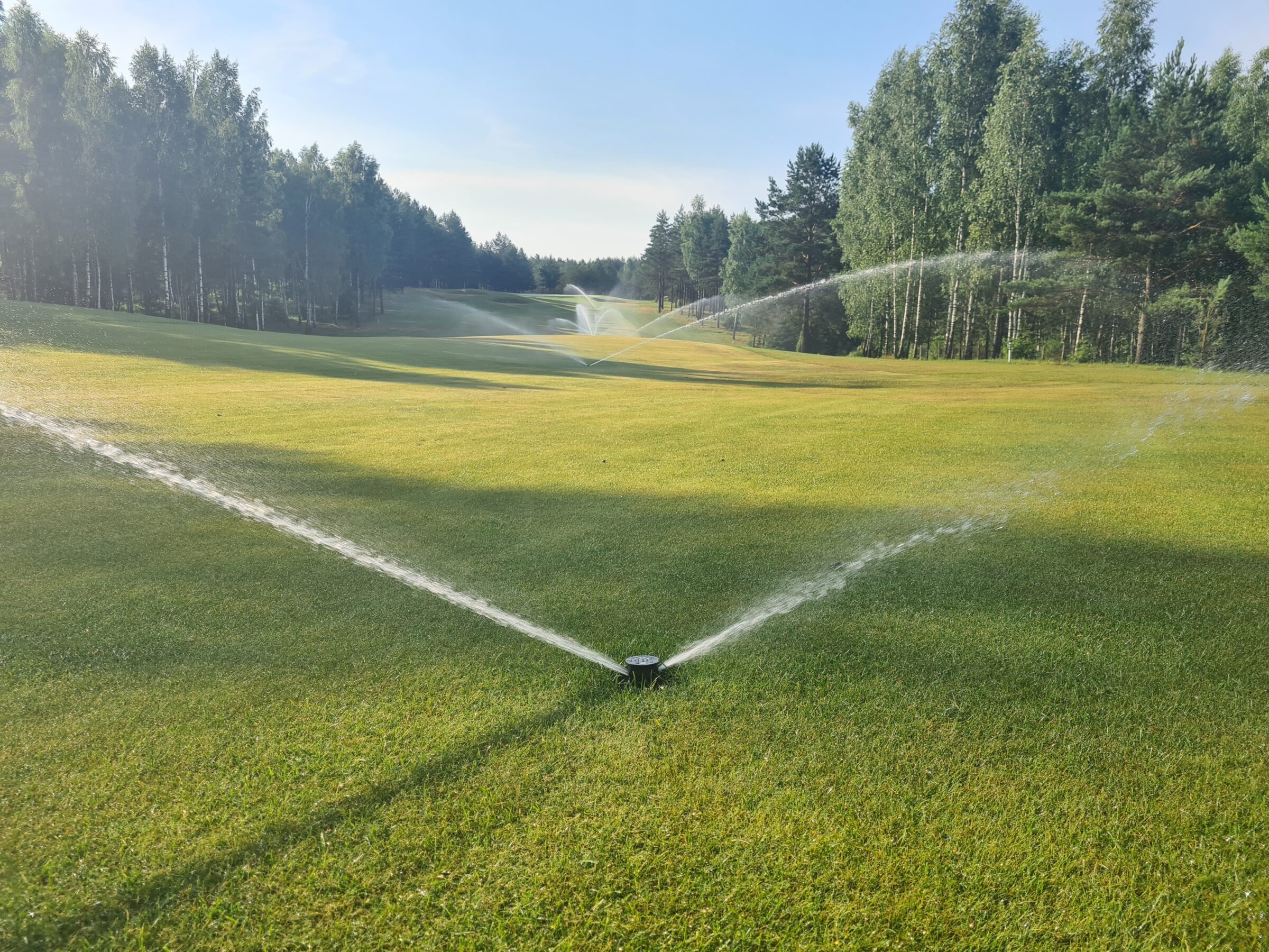 Lush green golf course with sprinklers watering the grass, surrounded by trees under a clear blue sky, creating a serene outdoor setting.