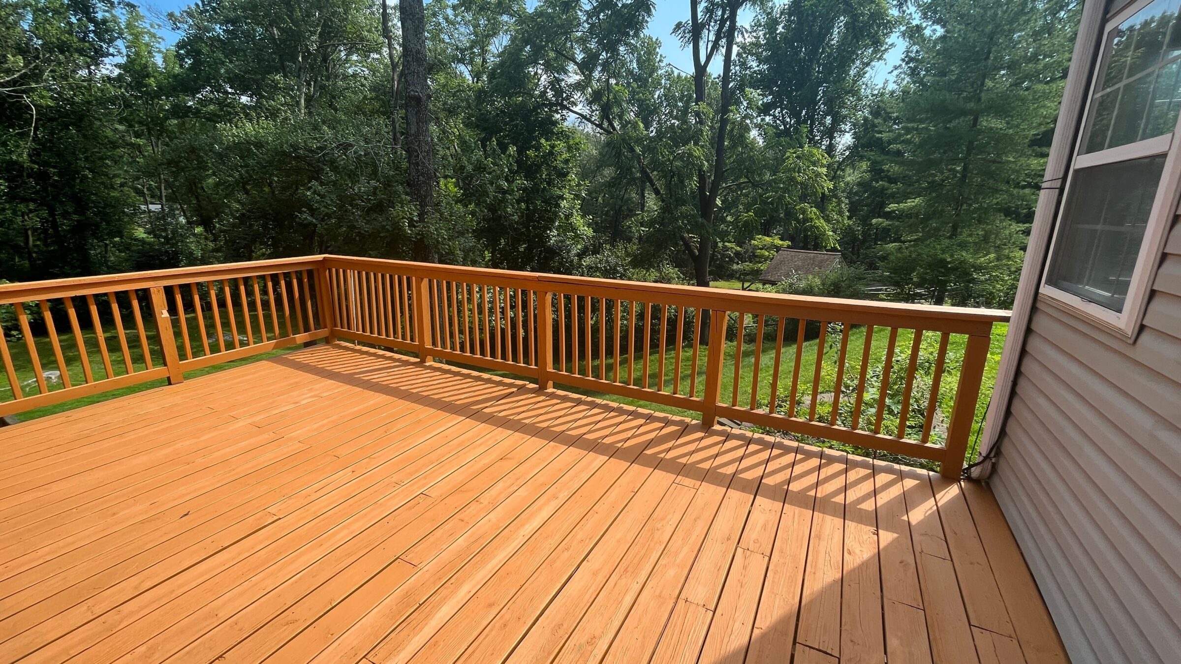 A wooden deck overlooks a lush green backyard surrounded by tall trees under a clear blue sky. No people or landmarks visible.