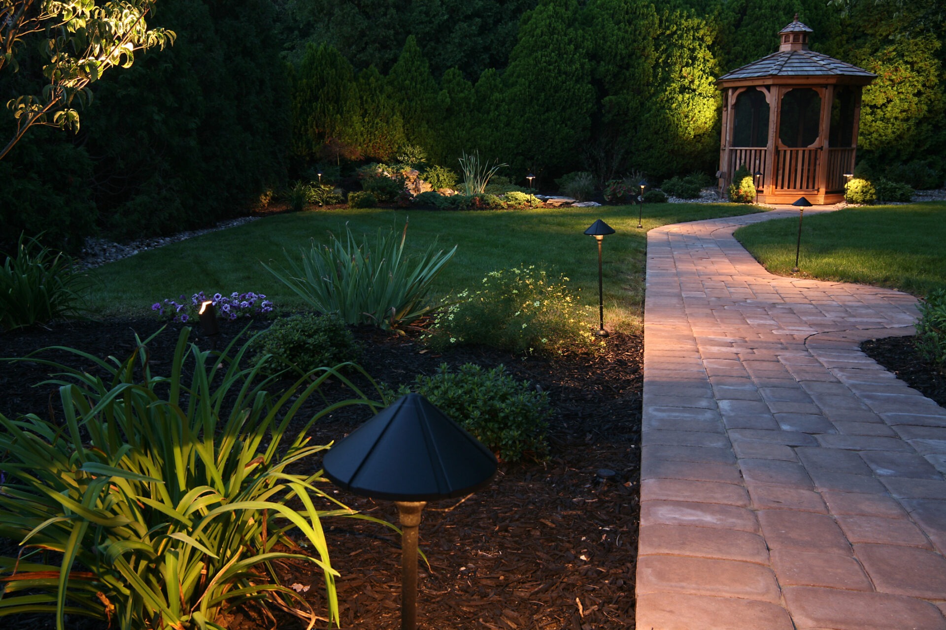 A beautifully lit garden path leads to a wooden gazebo, surrounded by lush greenery and garden lights illuminating the scene.