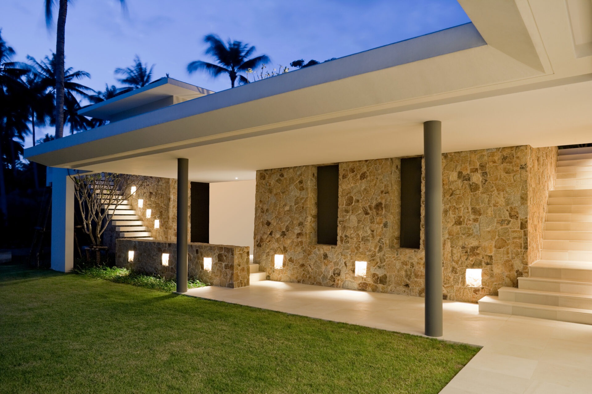 Modern stone house with illuminated steps and open design. Palm trees in the background, evening setting, contemporary architectural style.