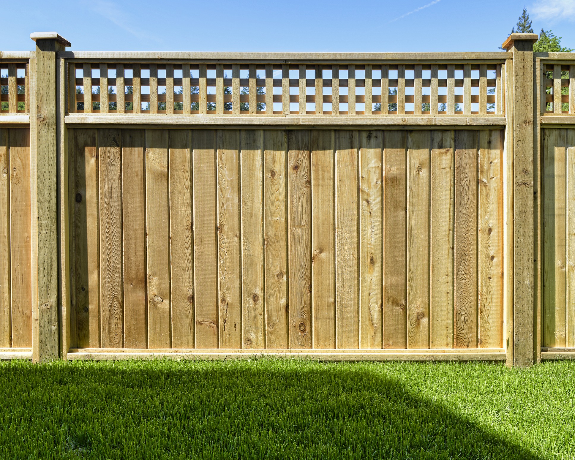 Wooden fence with lattice detail stands on a lush green lawn, under a clear blue sky. Trees are visible in the background.