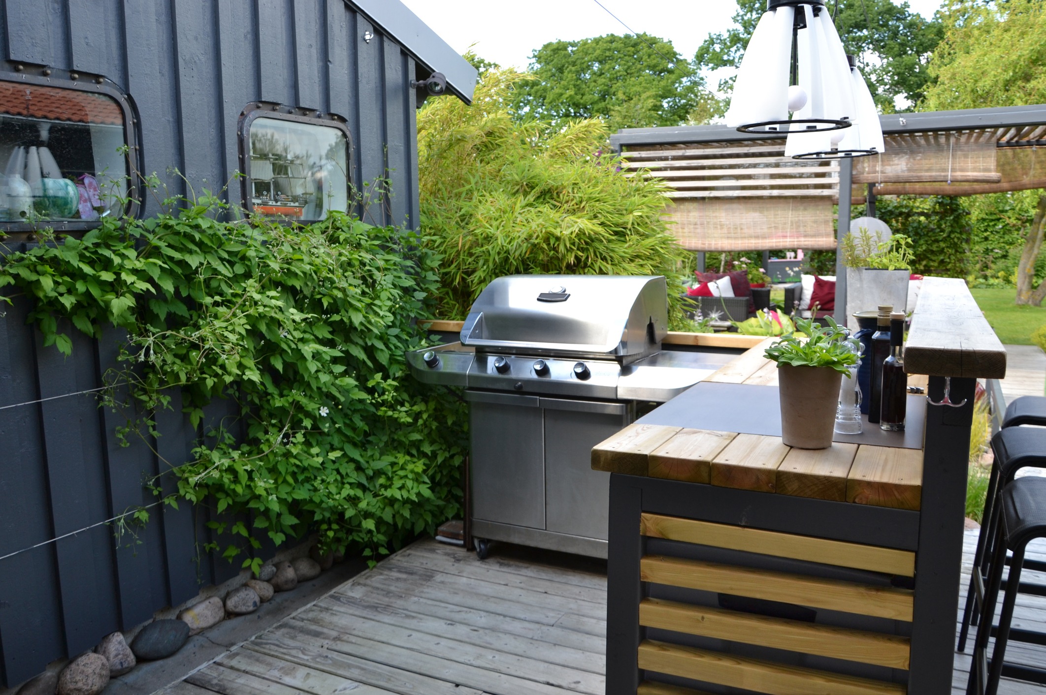 Outdoor kitchen featuring a stainless steel grill, wooden bar, and vibrant greenery on a patio. Cozy seating area visible in the background.