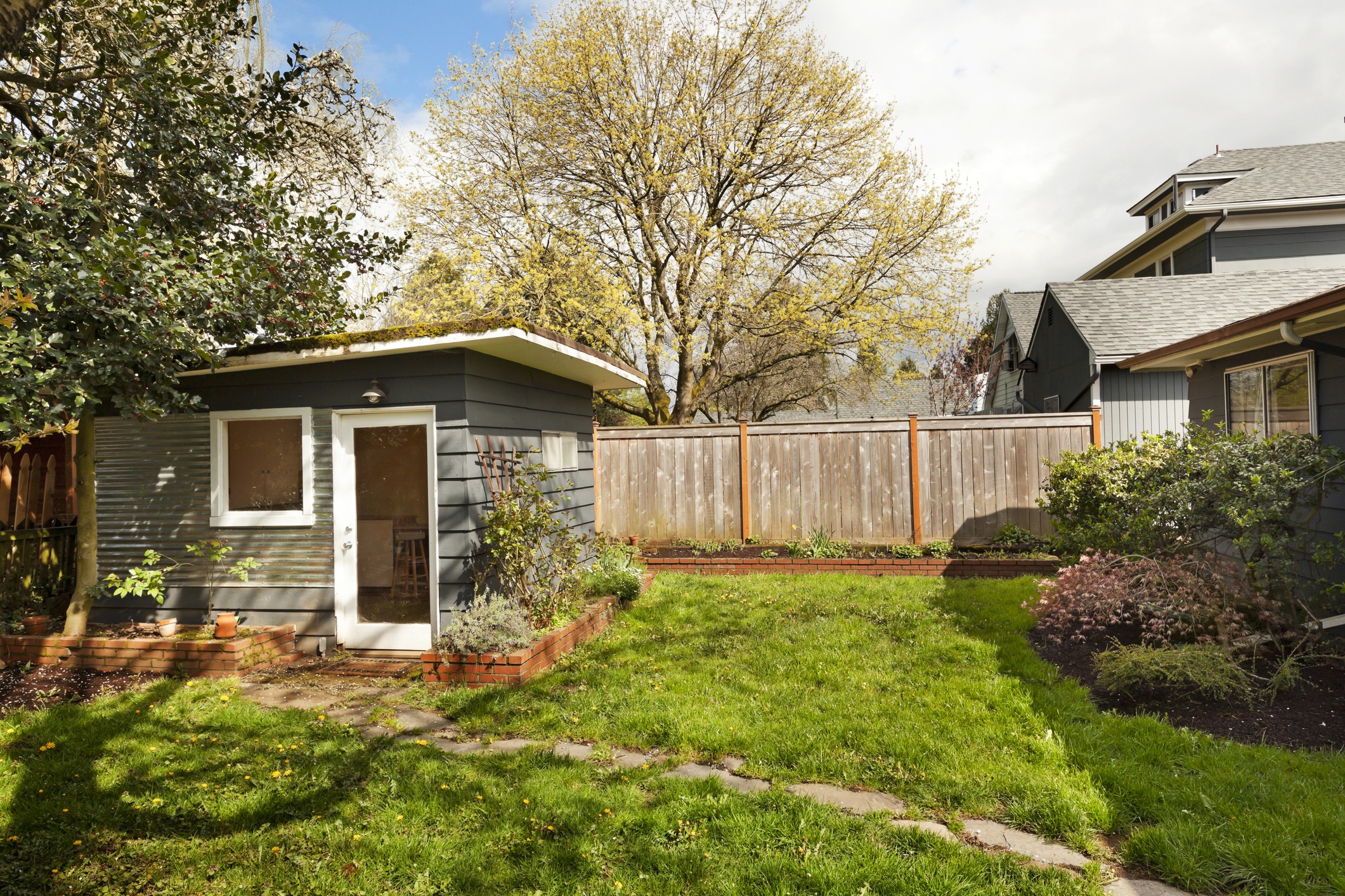 A small garden with a gray shed, vibrant green grass, and trees, surrounded by wooden fencing, under a partly cloudy sky.