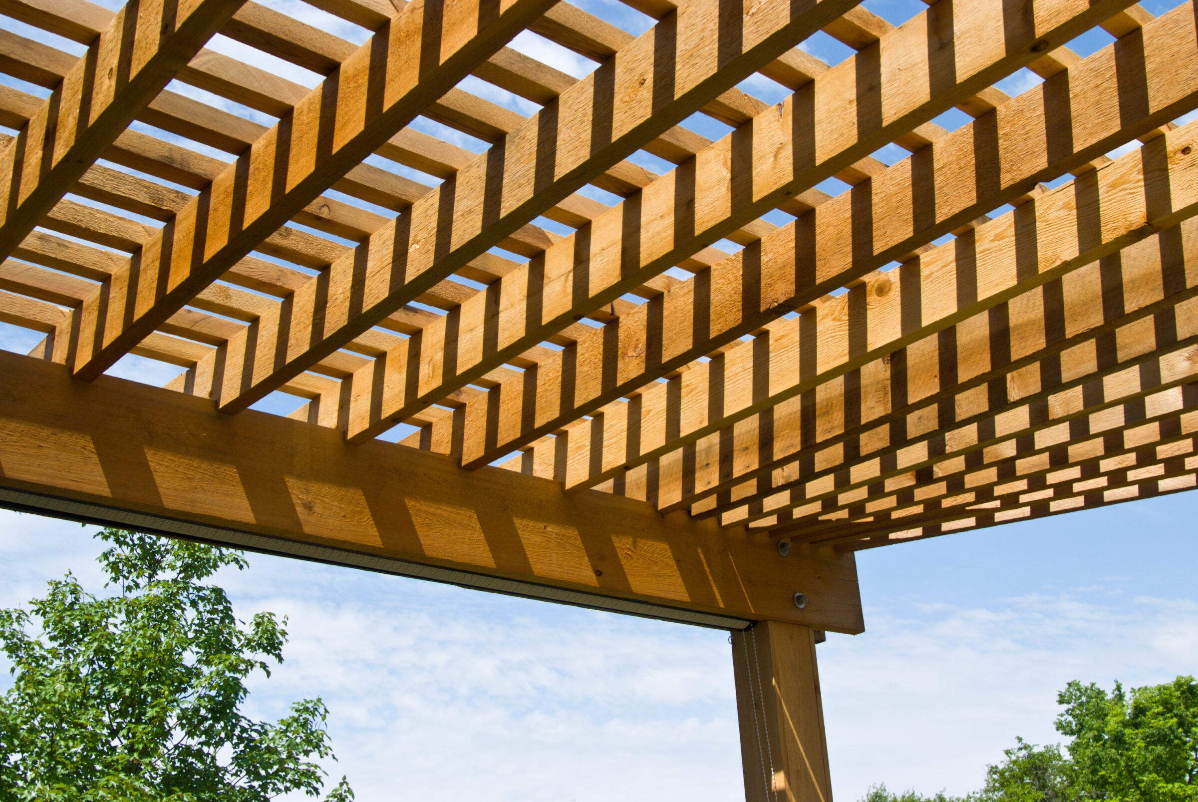 Wooden pergola casting geometric shadows against a clear blue sky with leafy green trees in the background, creating a serene outdoor atmosphere.