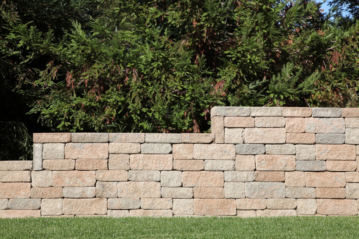 A stone retaining wall in a garden setting, surrounded by lush green trees and grass, under a clear sky.