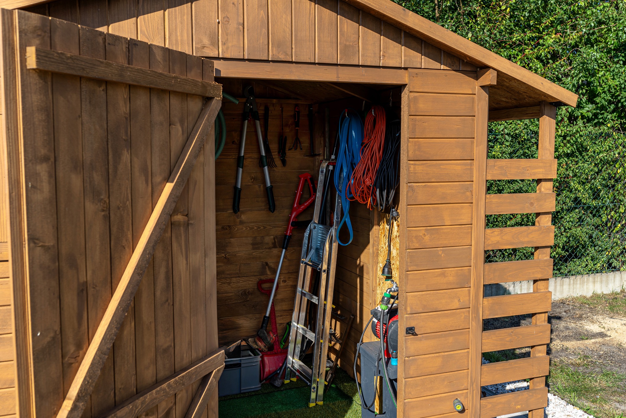 A wooden garden shed with open door displays various tools, a ladder, hoses, and equipment organized inside, surrounded by greenery and fencing.