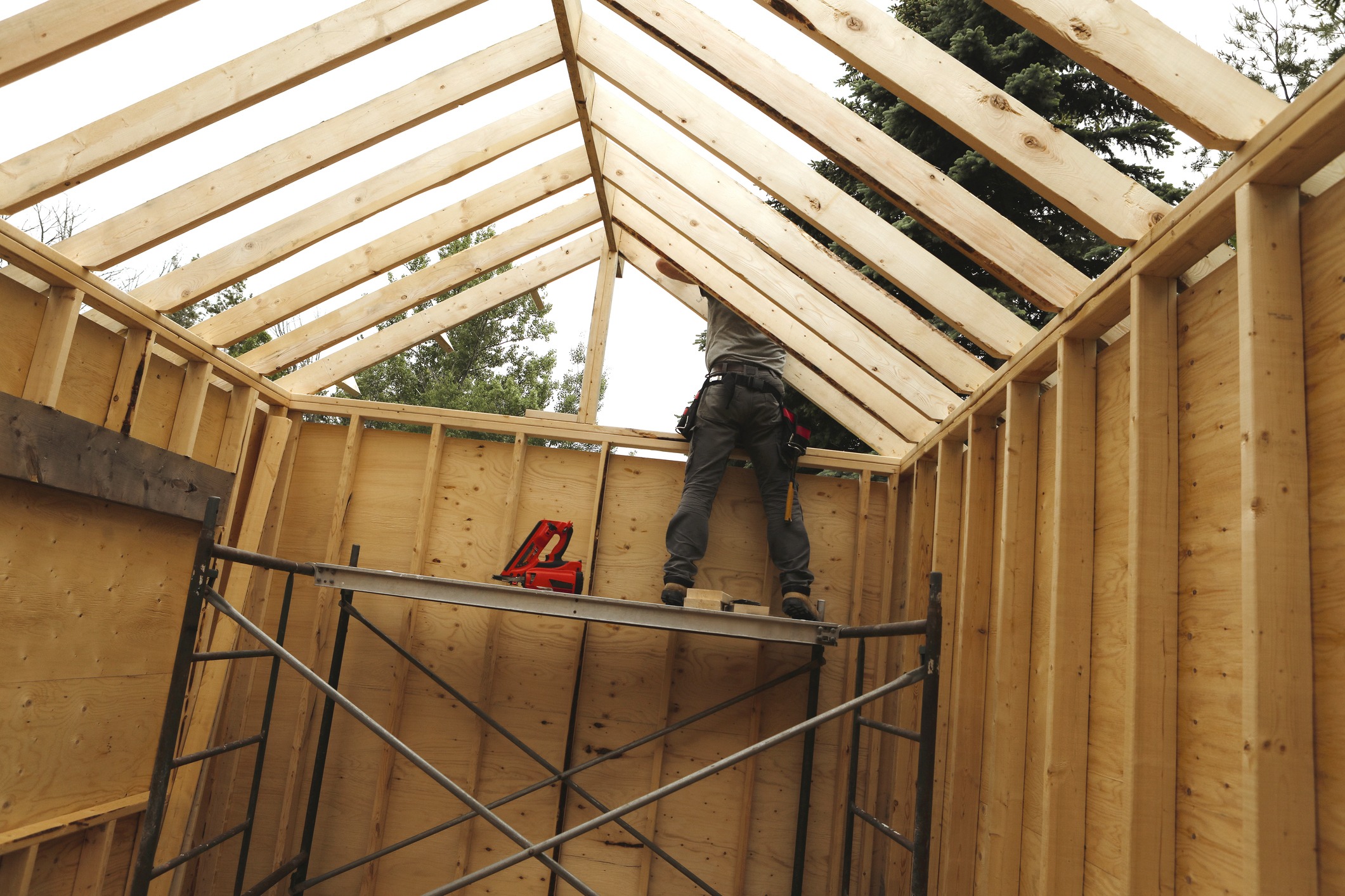 A person works on constructing a wooden roof frame on scaffolding, surrounded by trees, under a cloudy sky. Tools are visible nearby.