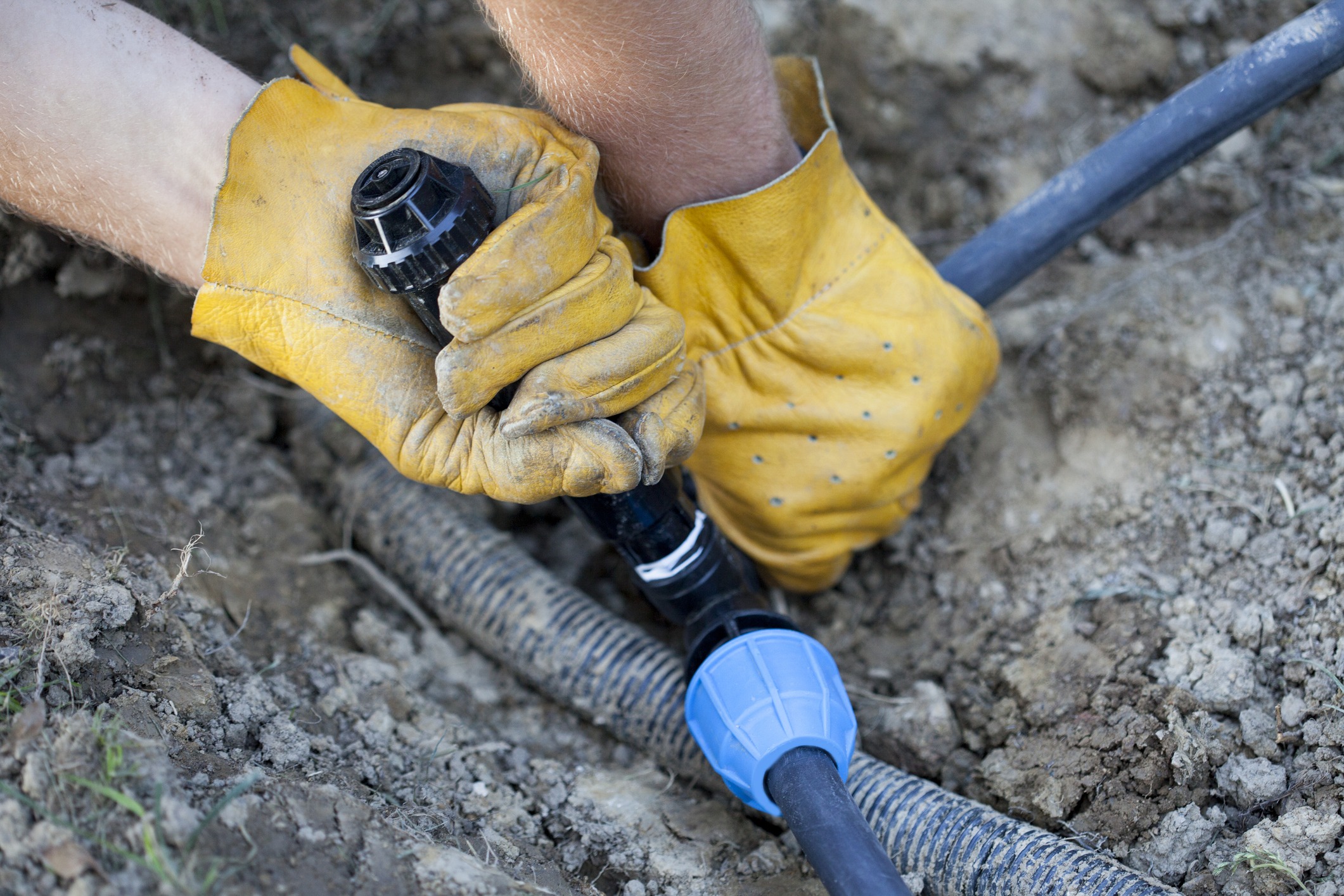 A person wearing yellow gloves connects irrigation pipes in a dirt-filled outdoor setting, focusing on plumbing or landscaping work.