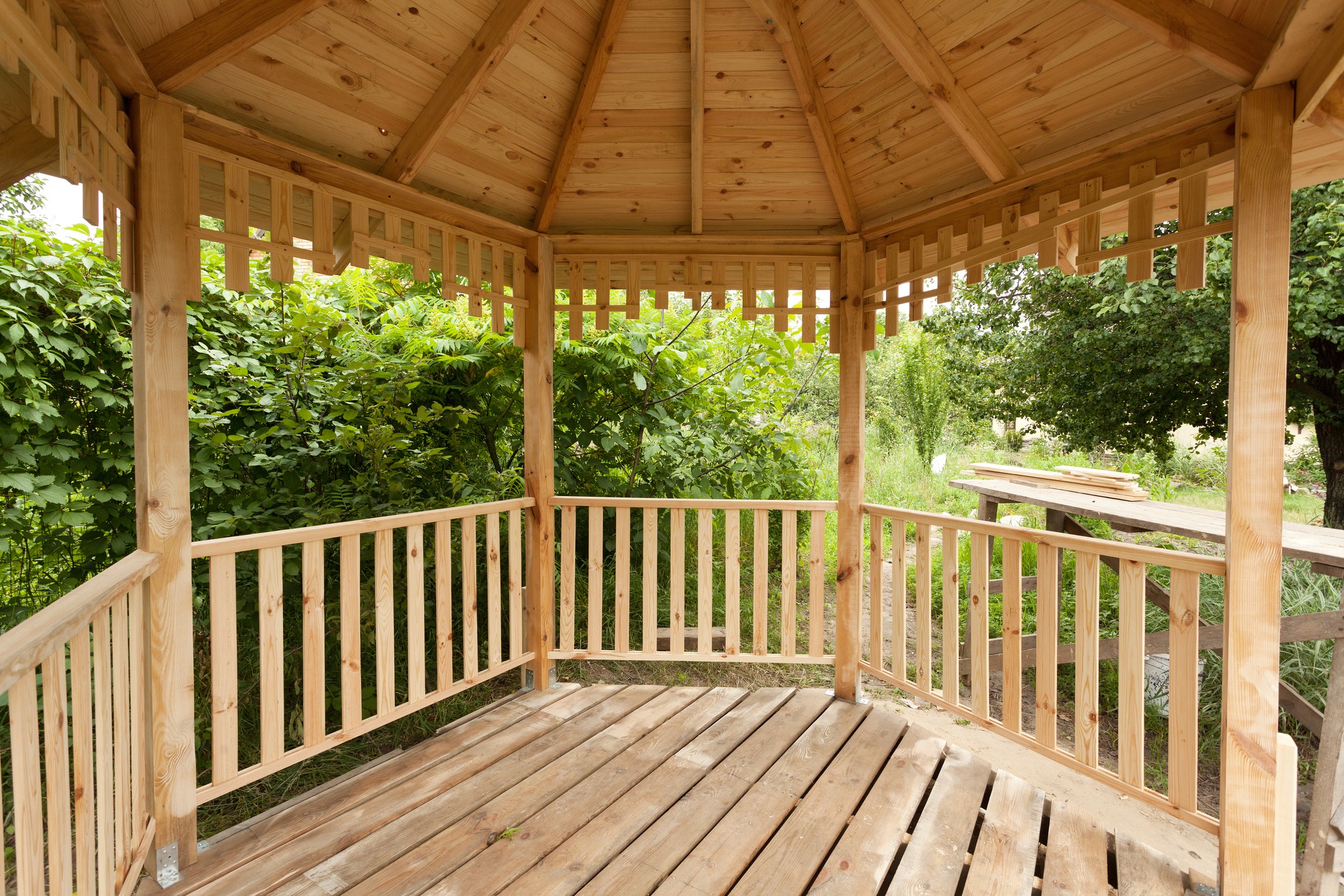 A wooden gazebo with a slatted floor and railings stands amidst lush green foliage, creating a serene and natural outdoor setting.