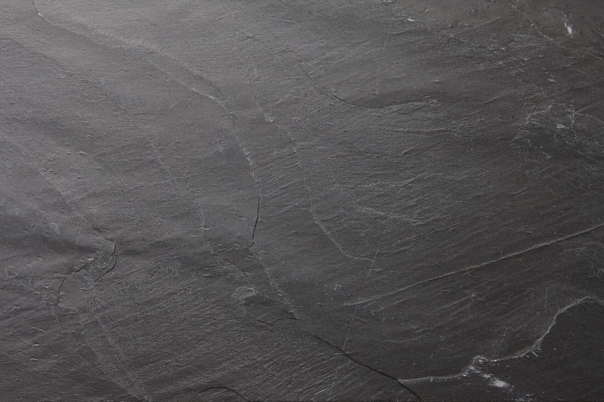 A close-up of a textured dark gray slate surface, displaying natural grooves and lines. The slate appears smooth yet rugged.