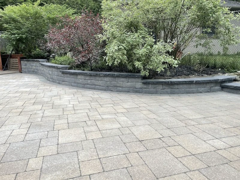 A spacious courtyard features a paved stone surface and a curved stone planter filled with lush green and red foliage against a fence.