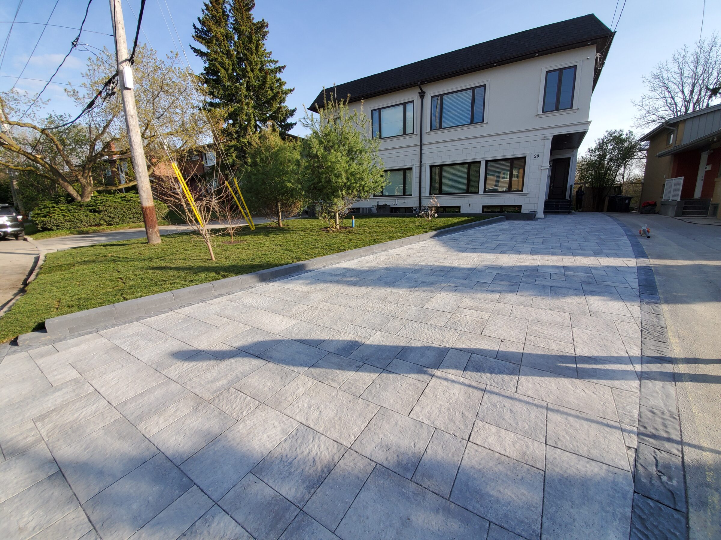 Modern two-story house with a freshly paved driveway, surrounded by trees and grass. Shadows of a person visible on the driveway.