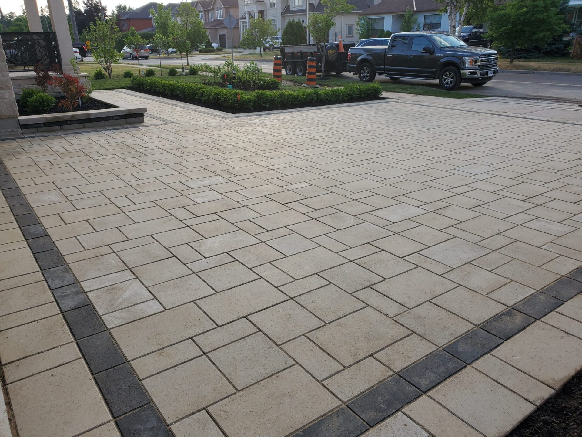 A driveway with neatly arranged paving stones, flanked by greenery; a parked truck is visible on a residential street.