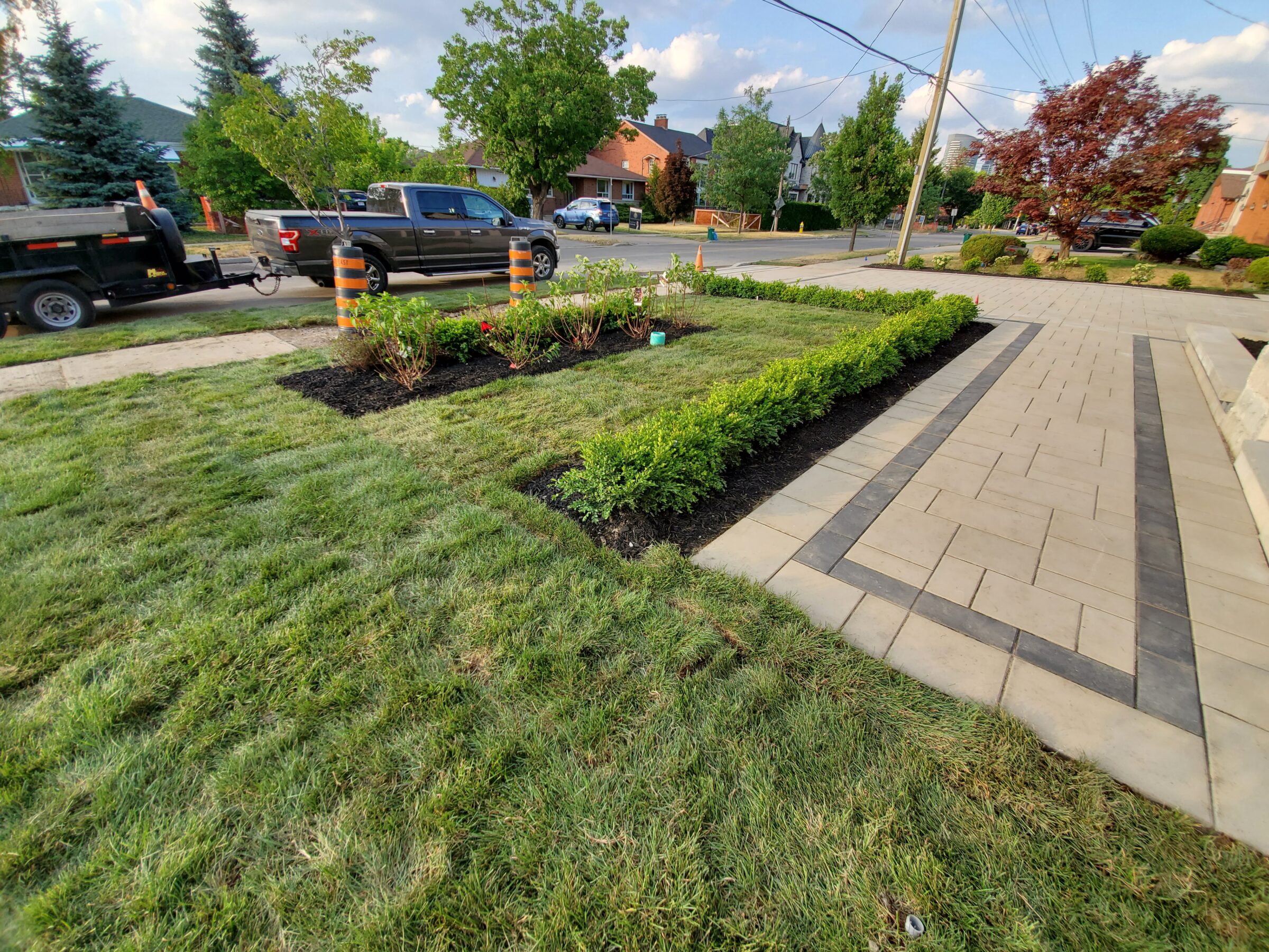 A neatly landscaped yard with hedges, a paved walkway, a parked truck with trailer, situated in a suburban neighborhood with trees and houses.