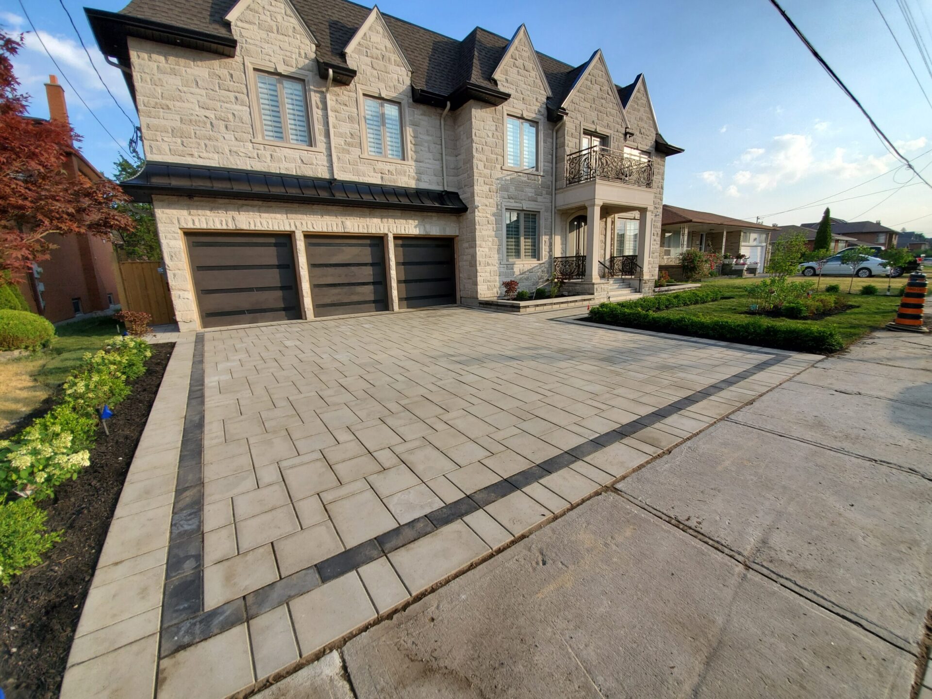 Modern stone house with three-car garage, landscaped garden, wide driveway, and neighboring houses. Clear sky and a single construction cone nearby.