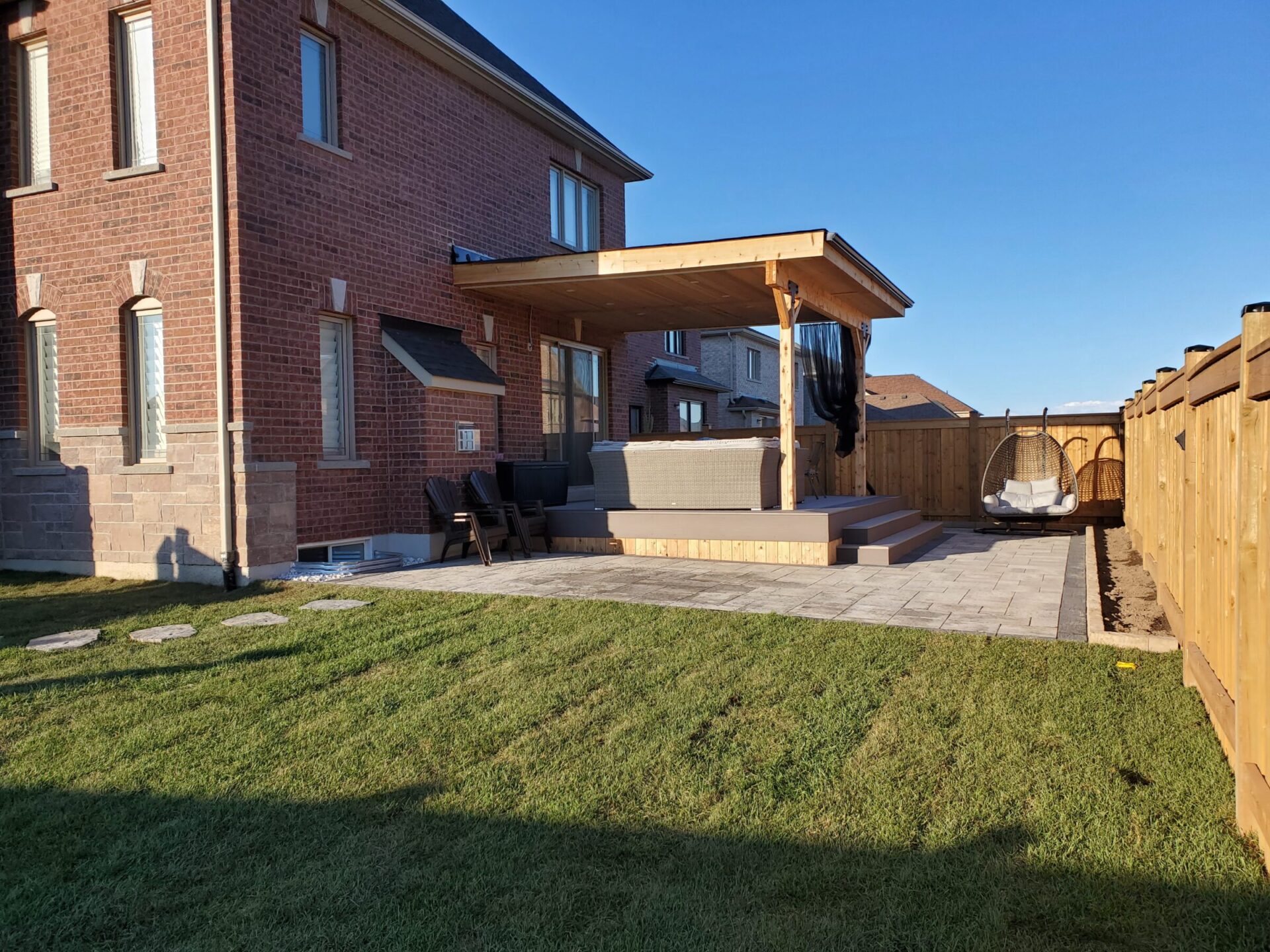Brick house with wooden pergola, patio furniture, and swing chair on a paved area. Well-maintained lawn and wooden fence surround the backyard.