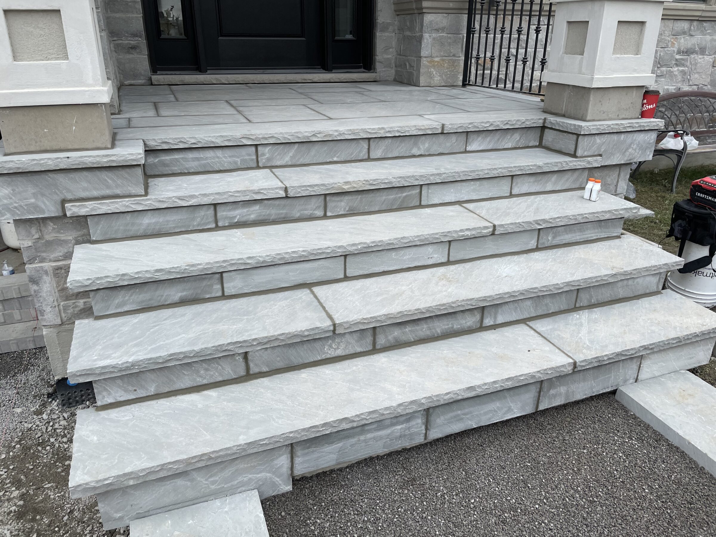 Stone steps leading to a porch with gray tiles. A small bench and construction equipment are visible nearby. Porch design is simple.