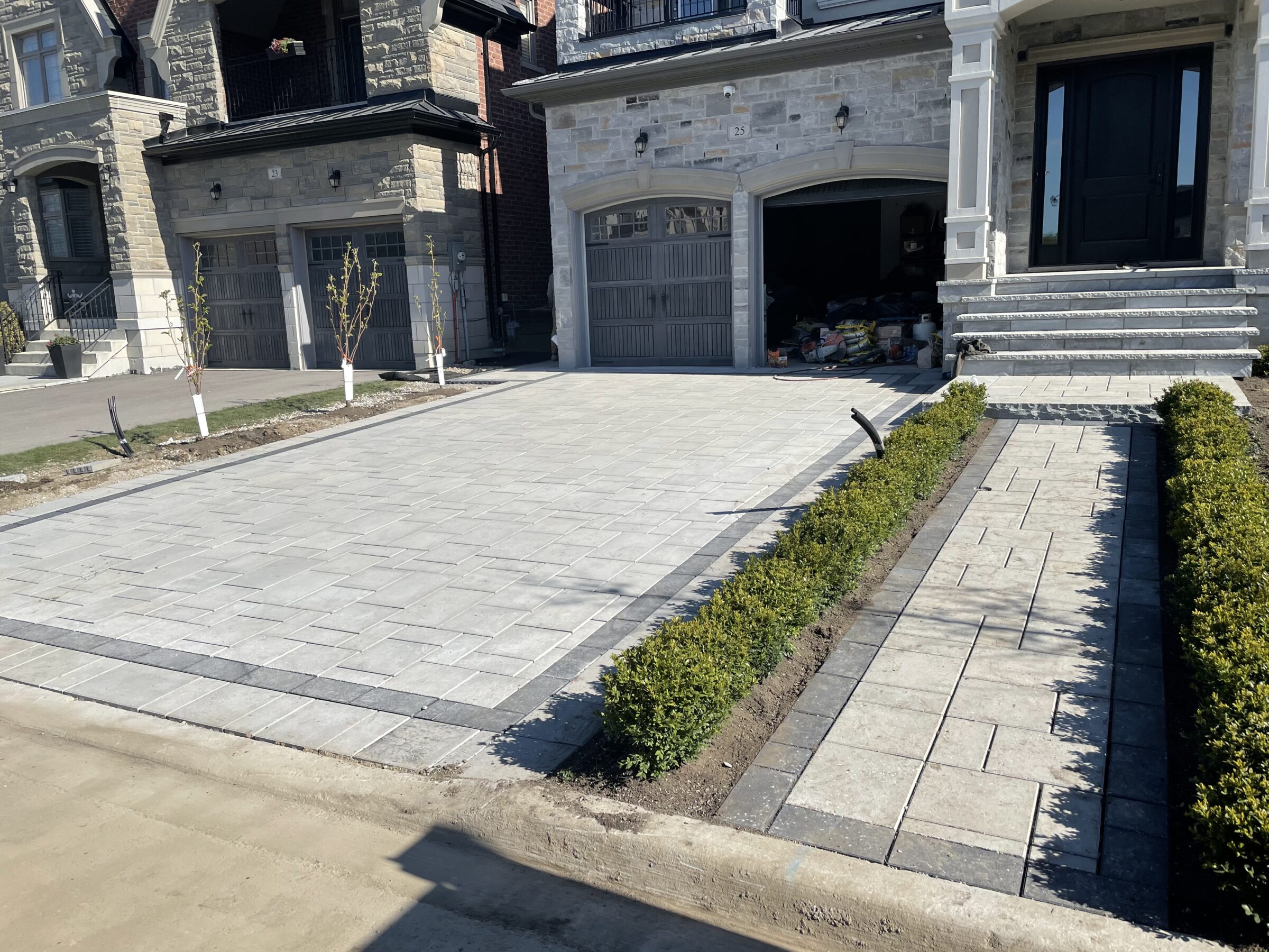 Driveway with light gray pavers leads to two modern garages. Small shrubs line the sidewalk. No people or recognizable landmarks are visible.
