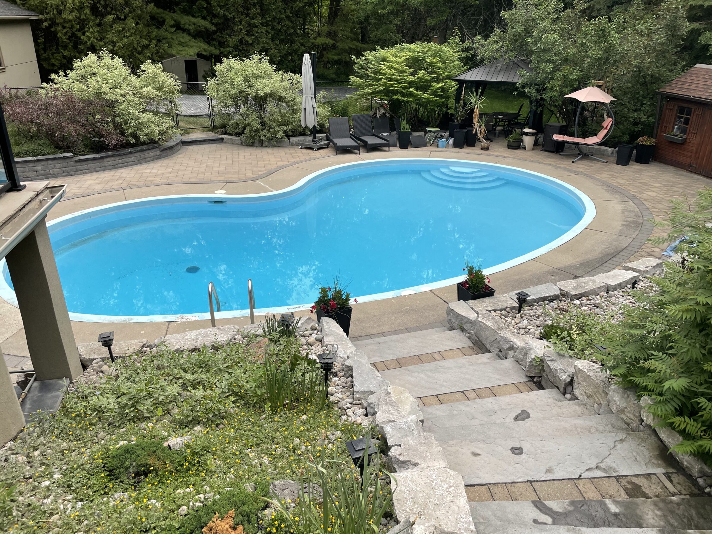 A serene backyard features a kidney-shaped pool, surrounded by greenery, lounge chairs, a gazebo, and a stone pathway leading to the pool area.