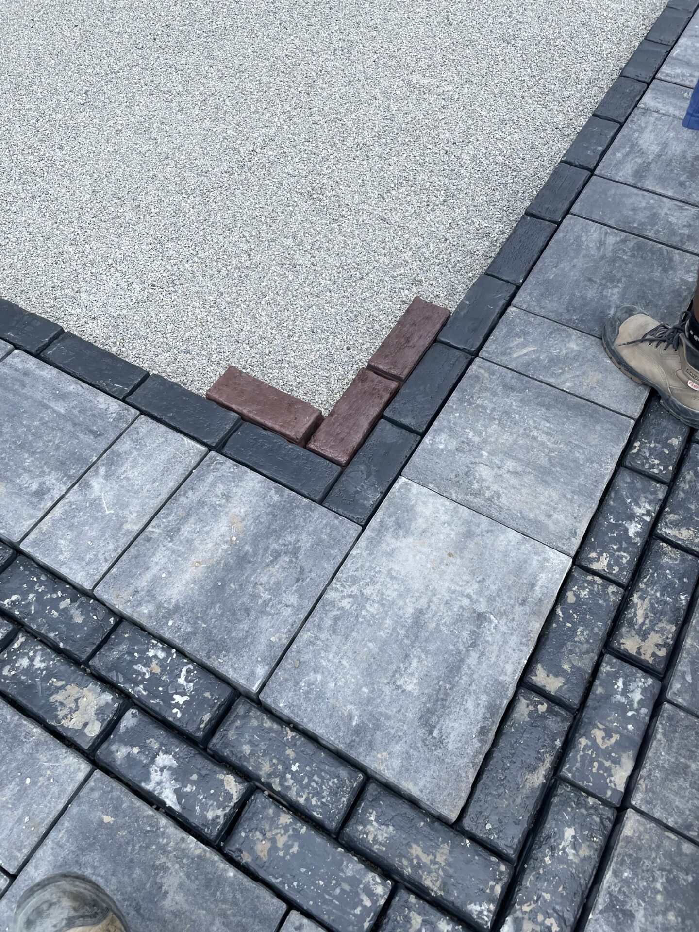 Paving project with gray bricks being laid, adjacent to exposed gravel. Visible person’s boot suggests ongoing construction work at the site.
