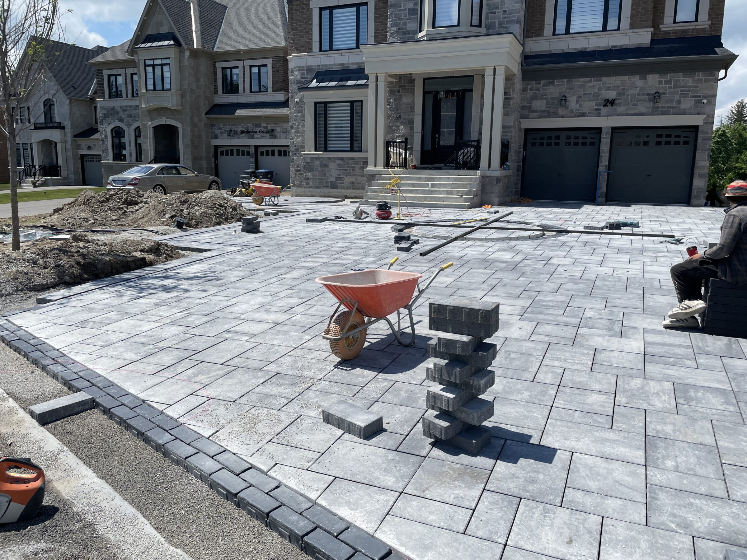 A person works on paving stones in front of a modern house. Tools and a wheelbarrow are visible, alongside another person seated.