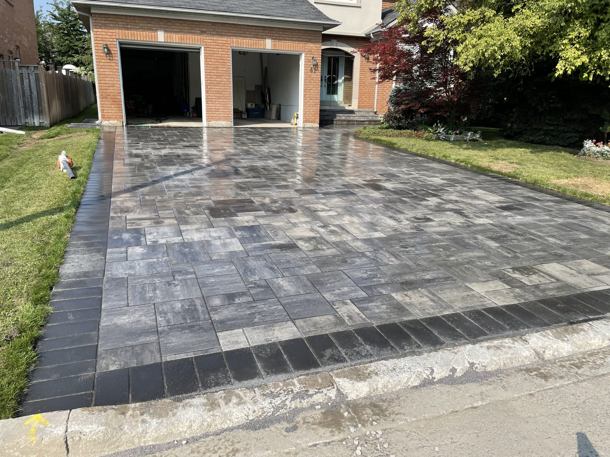 A newly paved stone driveway leads to a brick garage and house, with a person standing on adjacent grass, and trees surrounding.