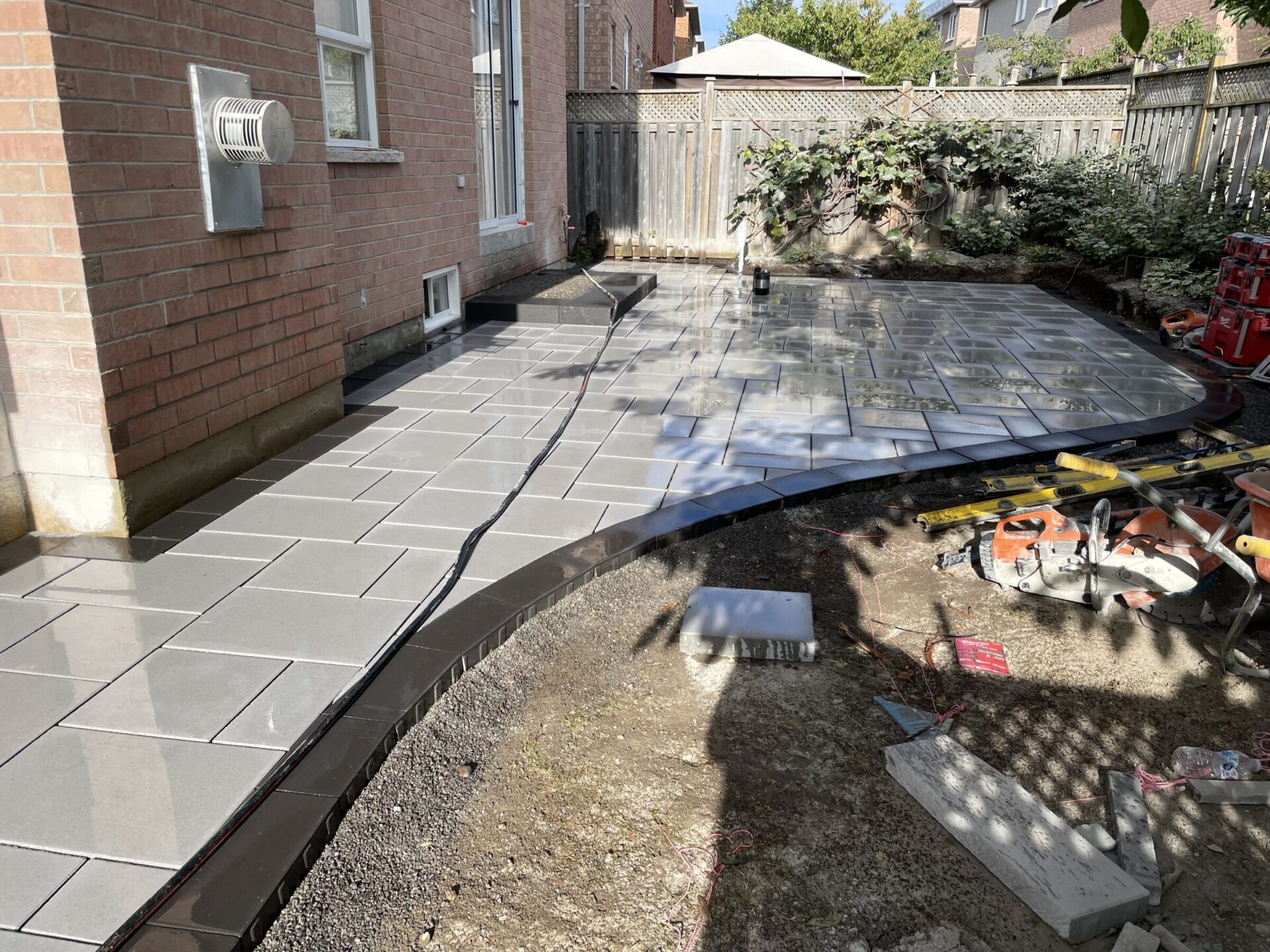 A newly laid patio with wet tiles, construction tools, and materials, near a brick house and wooden fence with scattered greenery.