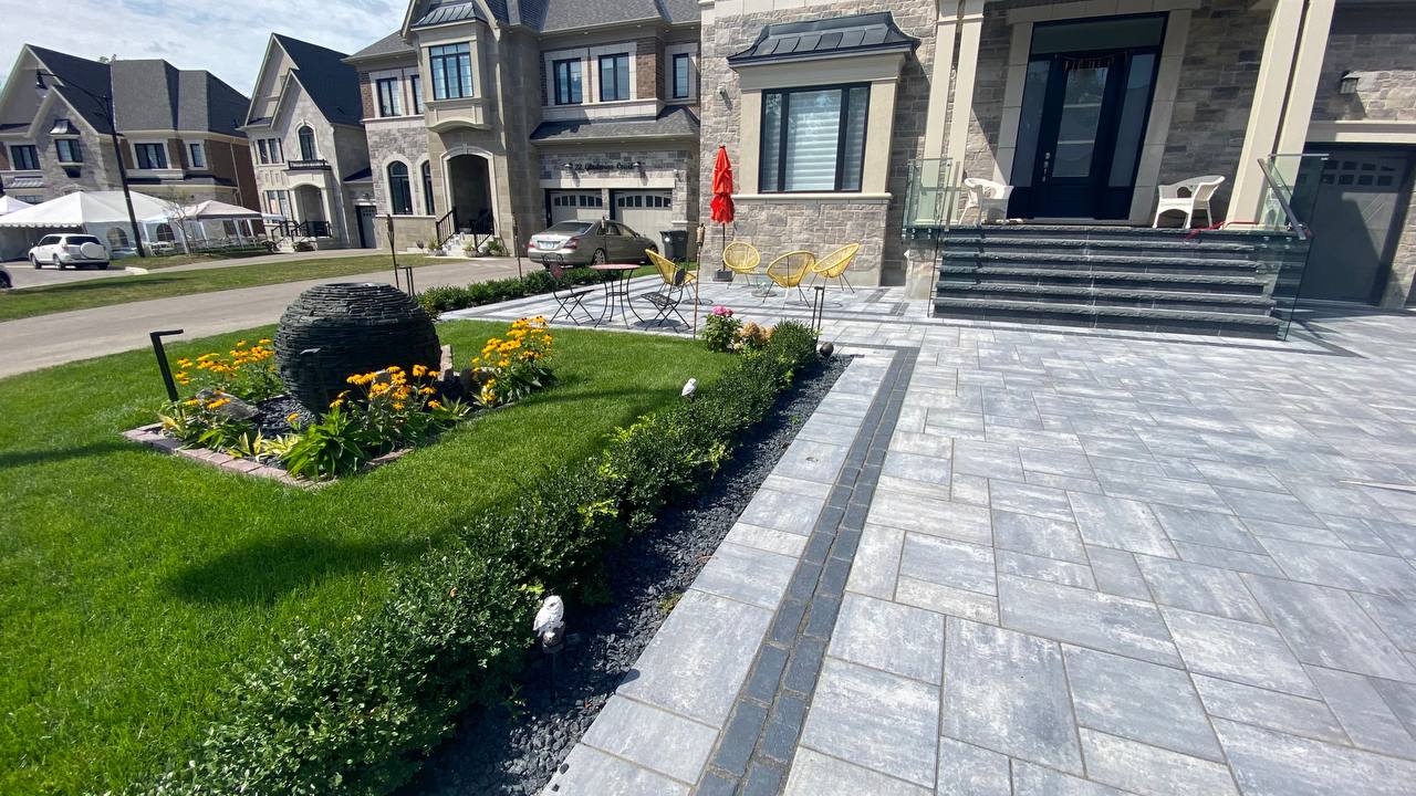 A modern suburban house with a manicured garden, patio furniture, stone pathway, and adjacent parked cars on a quiet residential street.