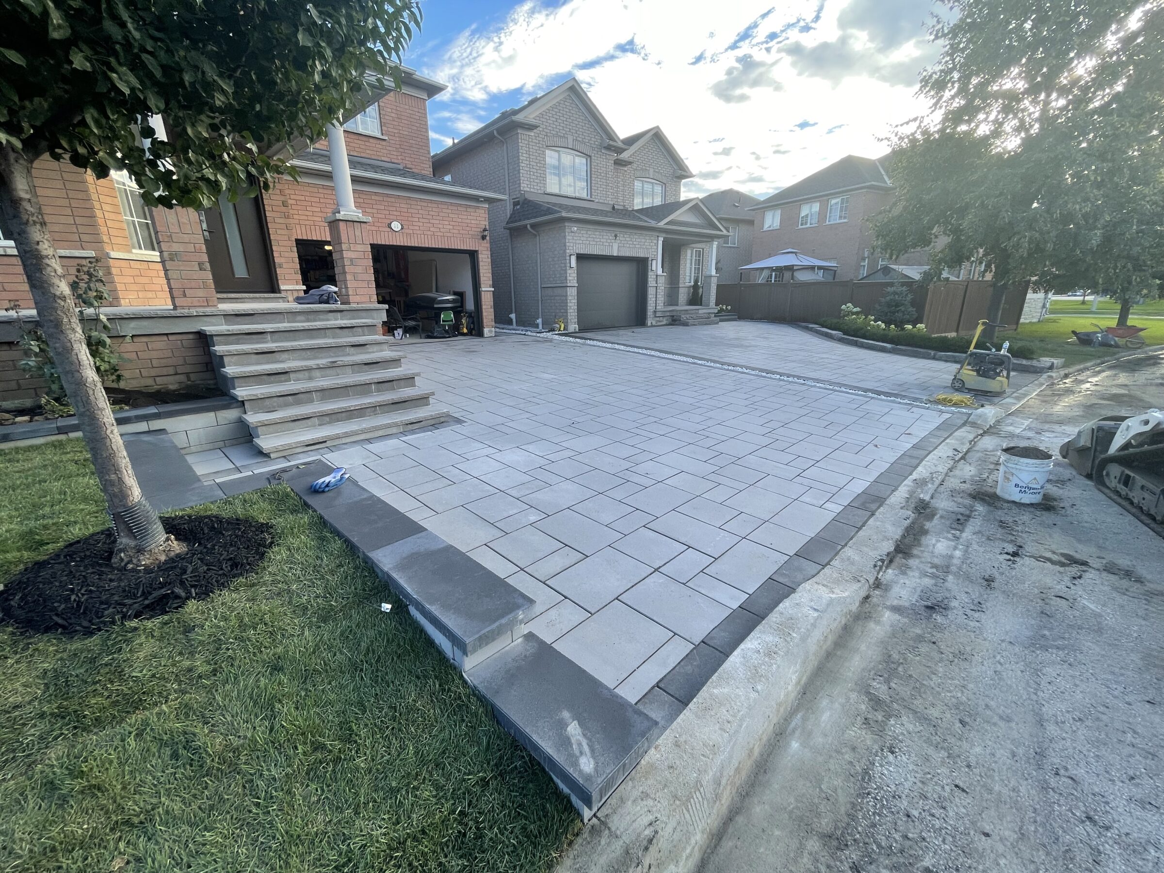 Suburban neighborhood scene with brick houses, a newly-paved driveway, stairs, trees, and a small lawn area. No visible people or landmarks.