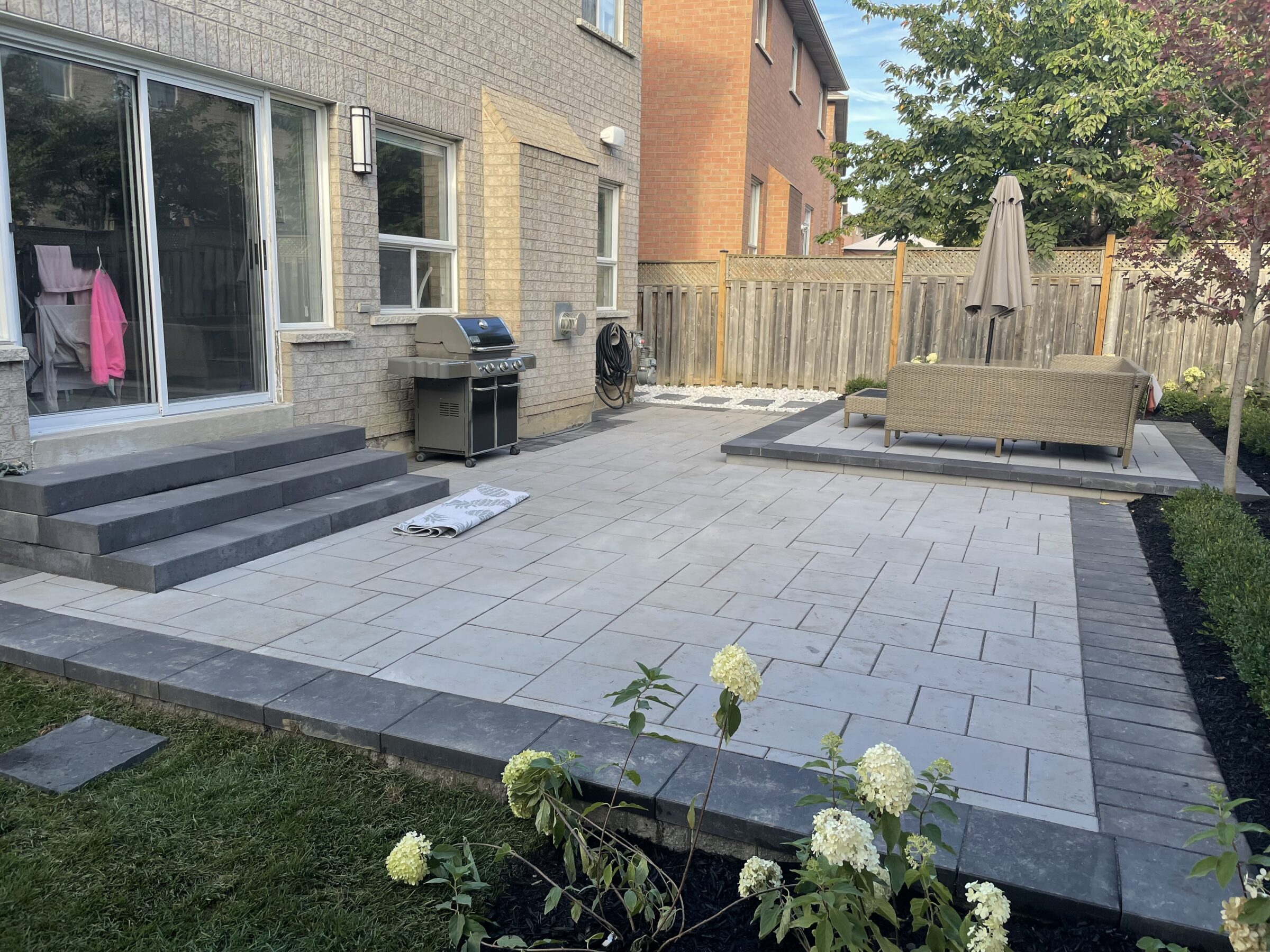 A modern backyard patio features a grill, potted plants, and an outdoor seating area with umbrella, surrounded by a wooden fence and garden.