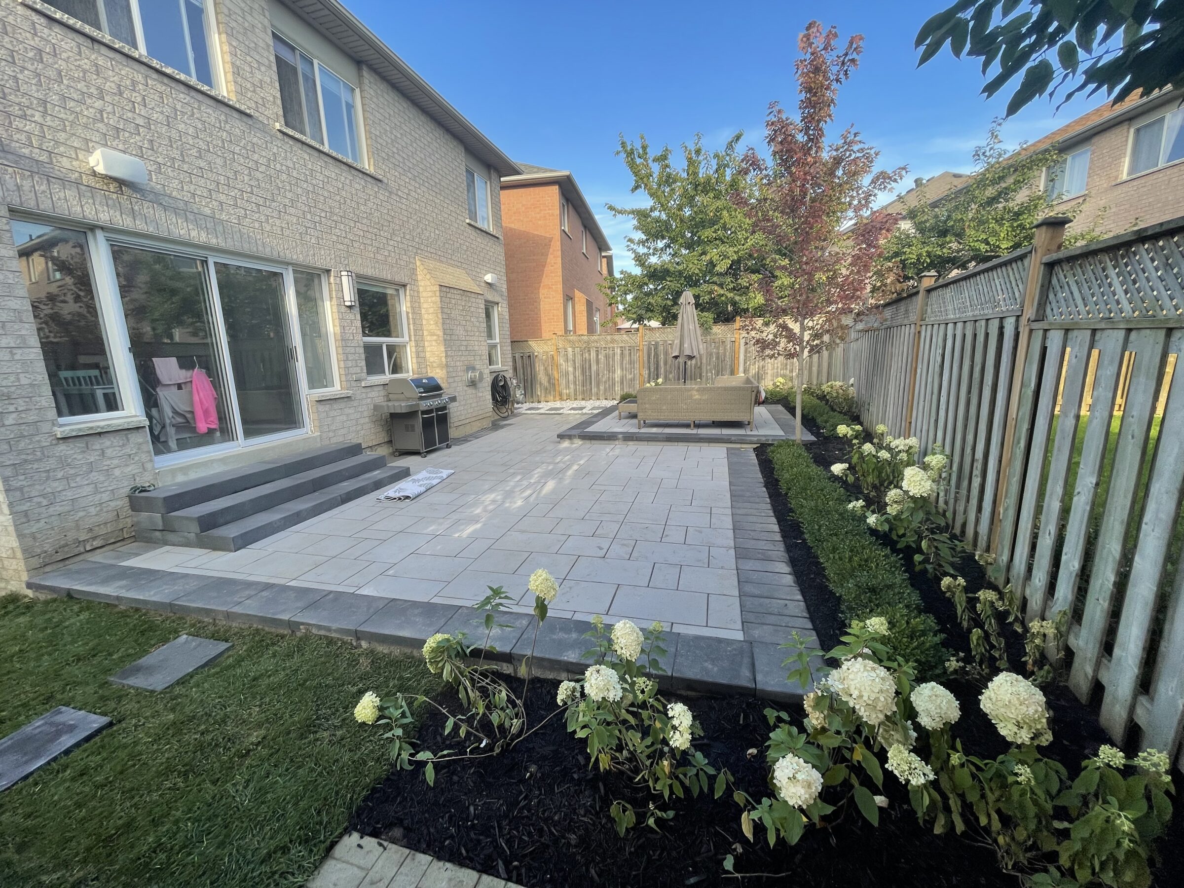 A well-maintained backyard features a paved patio, plants, outdoor seating, umbrella, grill, and a sliding glass door leading to the house.