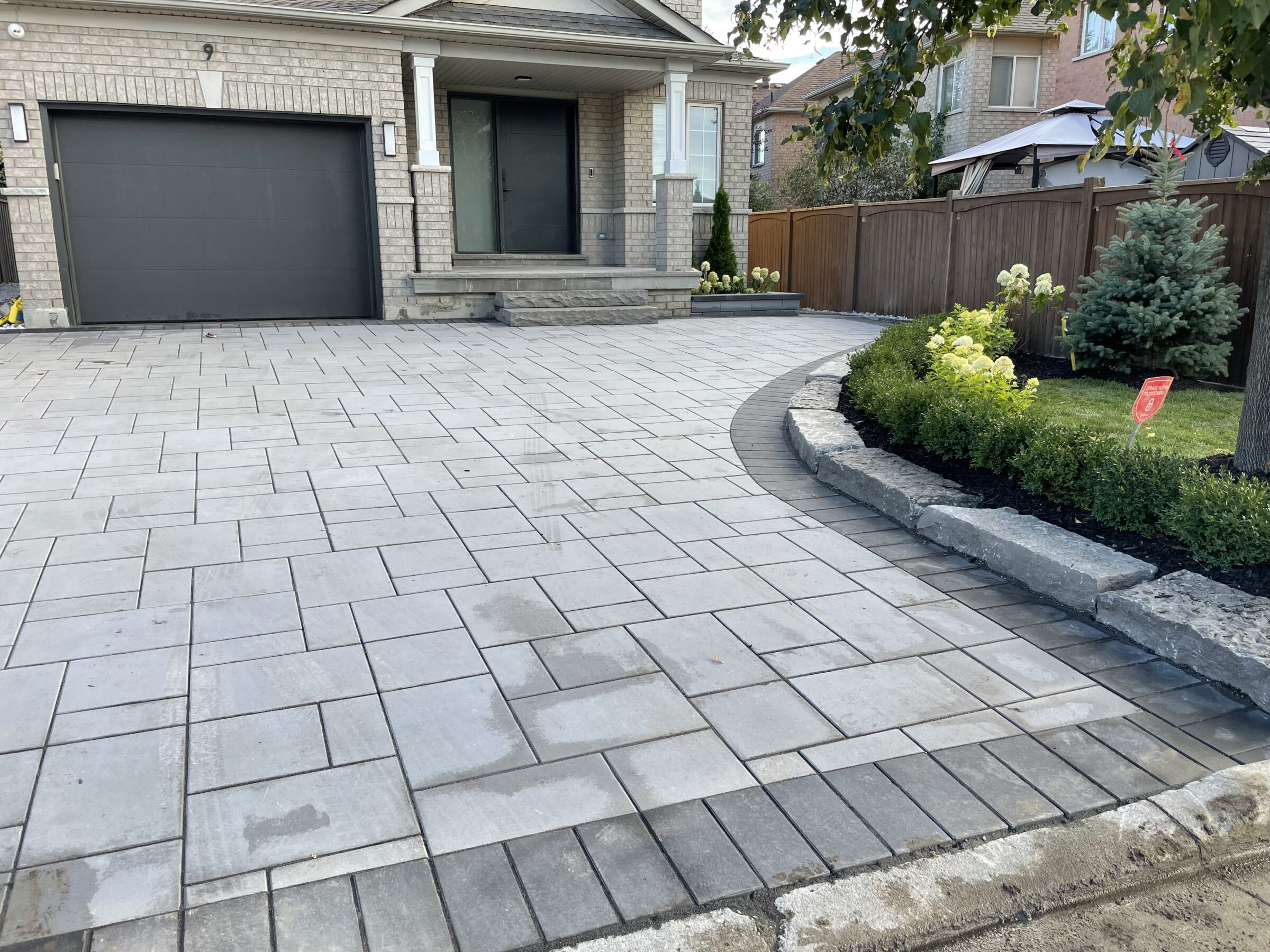 A modern house with a spacious paved driveway, garage, and landscaped garden featuring shrubs and a small tree, surrounded by a wooden fence.