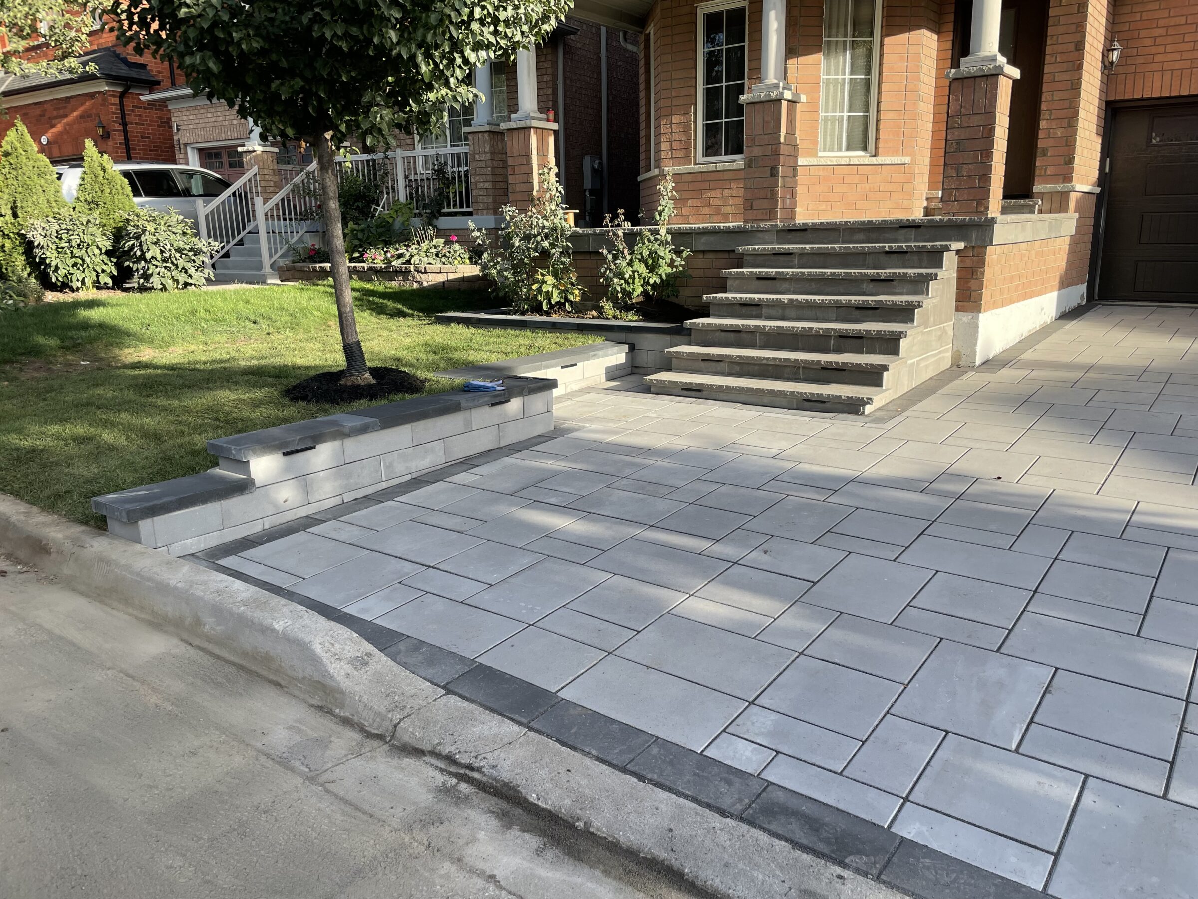Brick house with tiled driveway, steps, and landscaped garden. Tree and shrubs enhance the entrance. Sunny, residential suburban neighborhood setting.