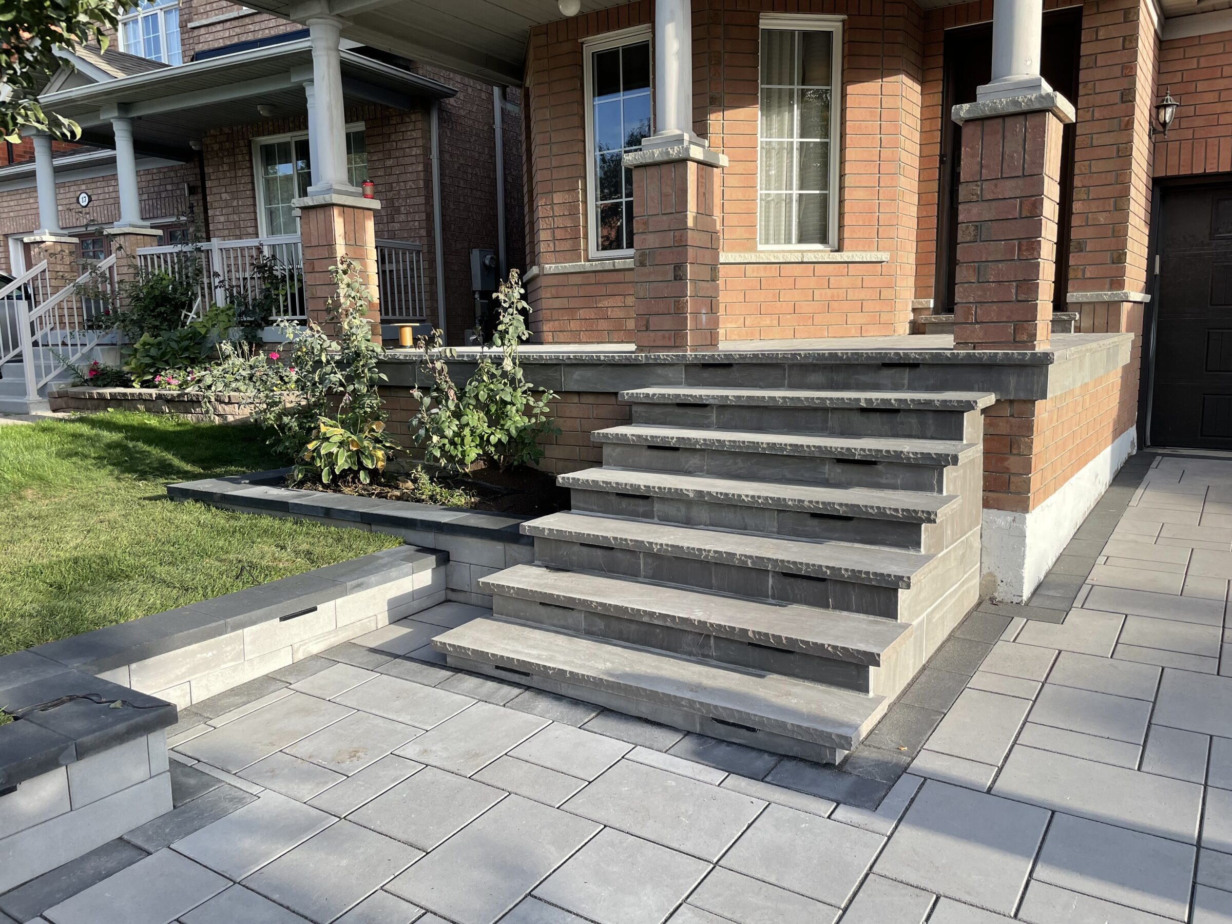 A red-brick house entrance with gray stone steps, small garden, and tiled walkway; porch supported by white columns.