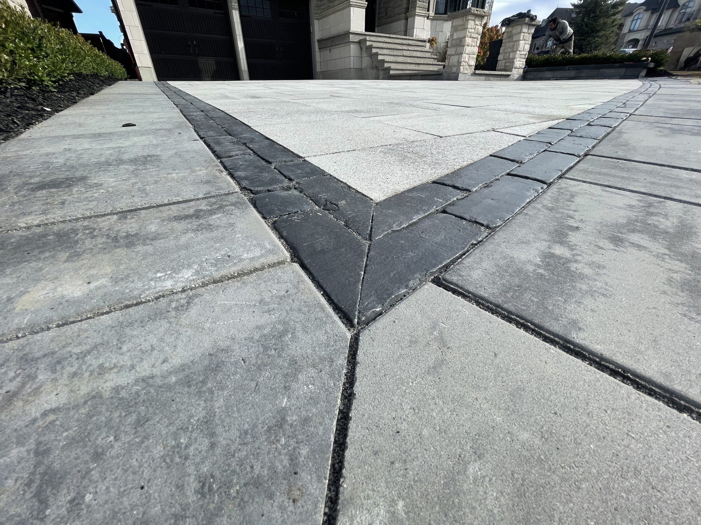 Close-up view of a patterned concrete driveway leading to a house entrance. Bushes and a person in the background add detail.