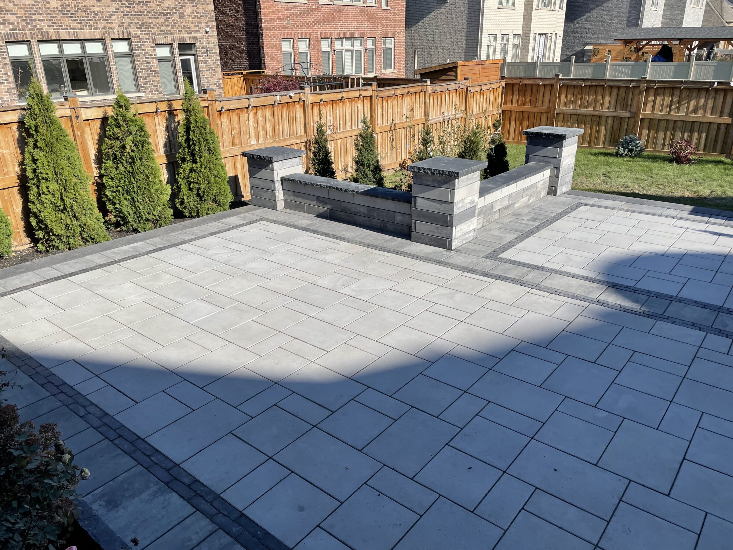 A well-paved backyard patio with surrounding evergreen trees and wooden fencing, bordered by residential brick and stone houses.