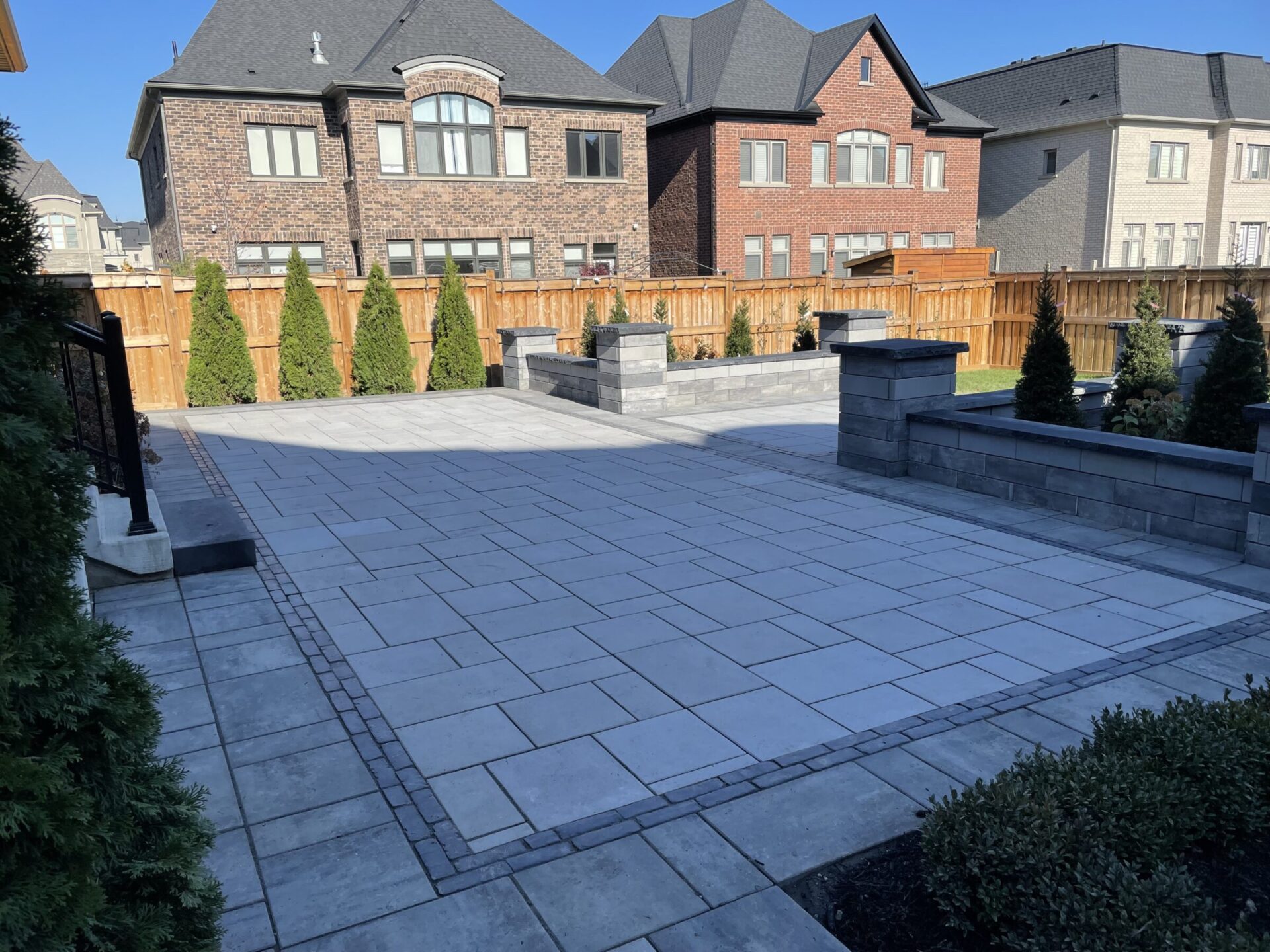 Backyard patio with stone tiles, brick houses, wooden fence, and rows of tall shrubs. Clear sky and modern residential architecture complete the scene.