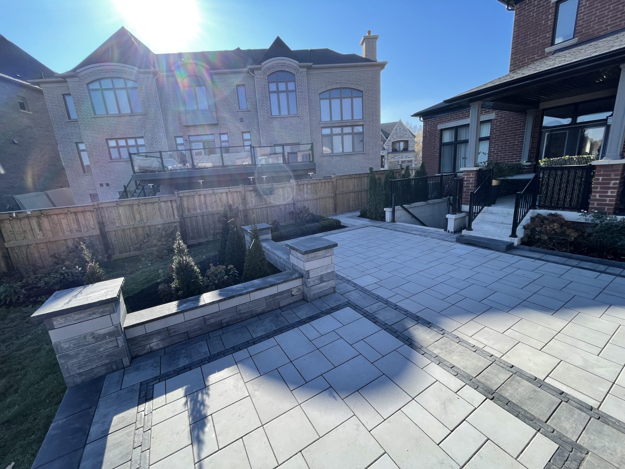 A sunny courtyard with paved tiles, surrounded by brick houses and a wooden fence, featuring minimal landscaping and modern architecture.