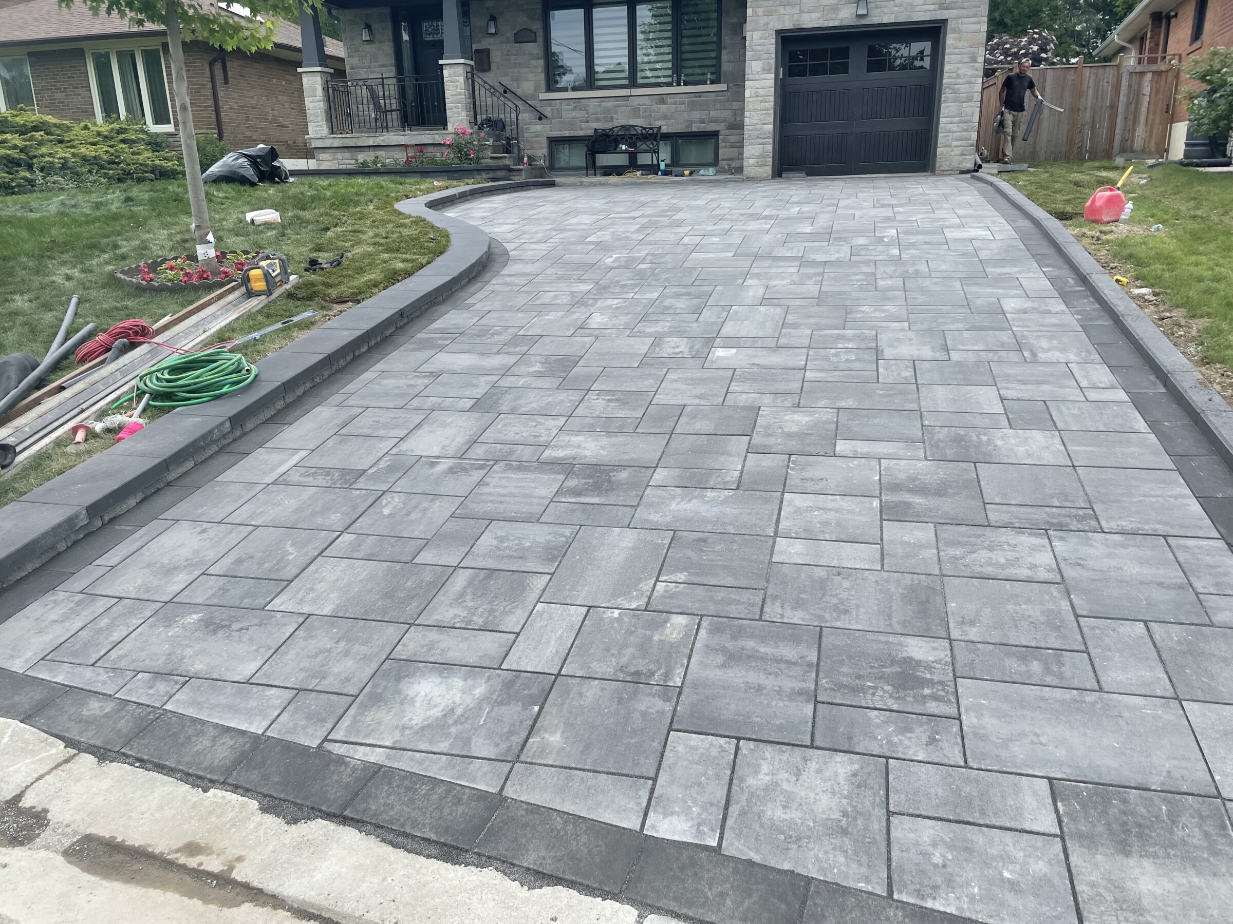 A newly paved driveway leads to a house with a person nearby. Garden tools and a hose are visible on the lawn.