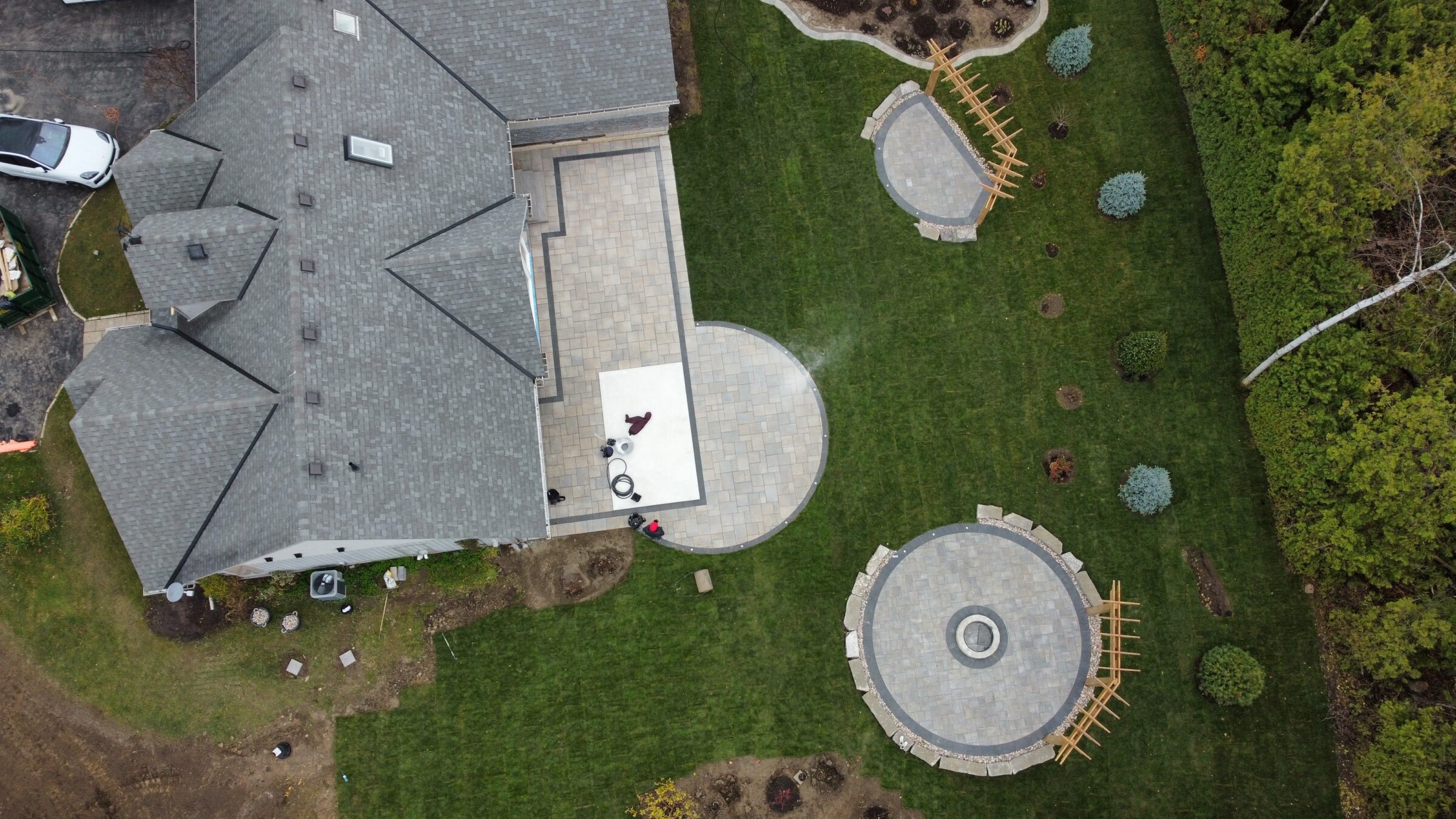 Aerial view of a house with gray roof, landscaped backyard, circular stone patios, green grass, and two people on the patio area.