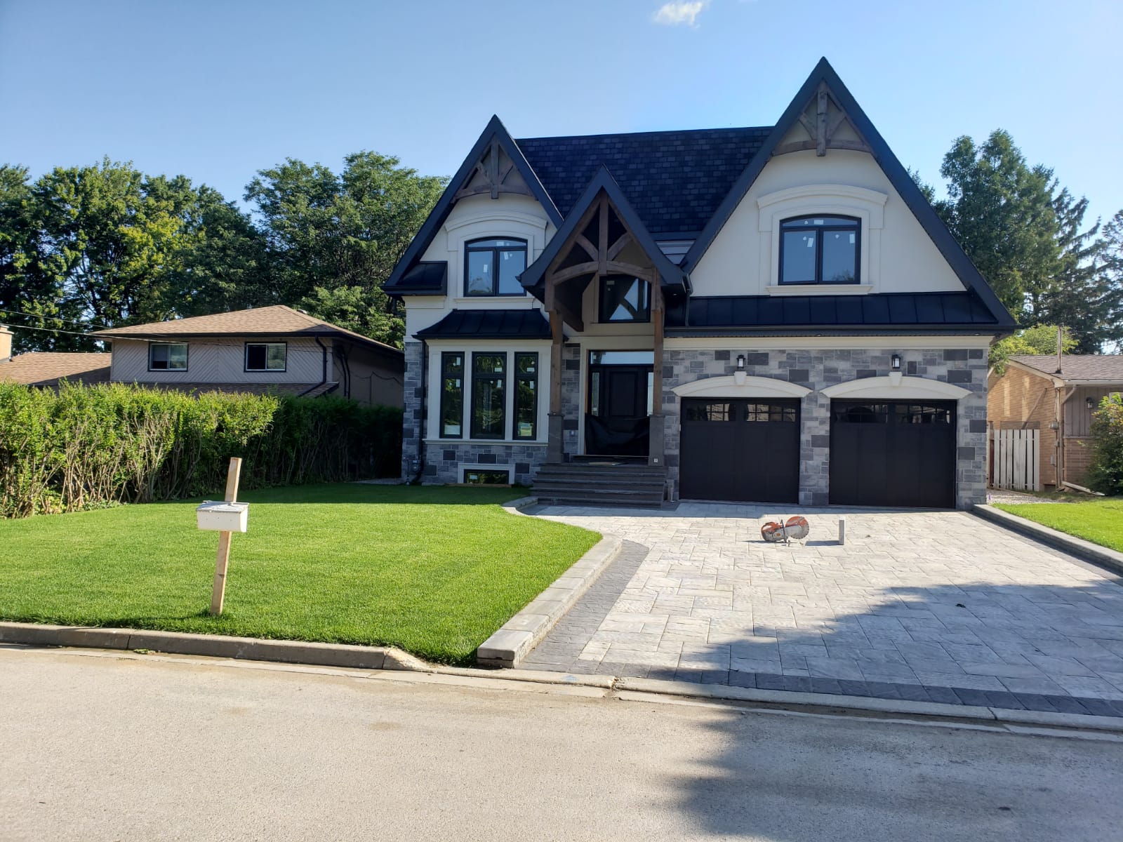 A modern, two-story house with a steep roof and large driveway, surrounded by greenery and neighboring homes, under a clear blue sky.
