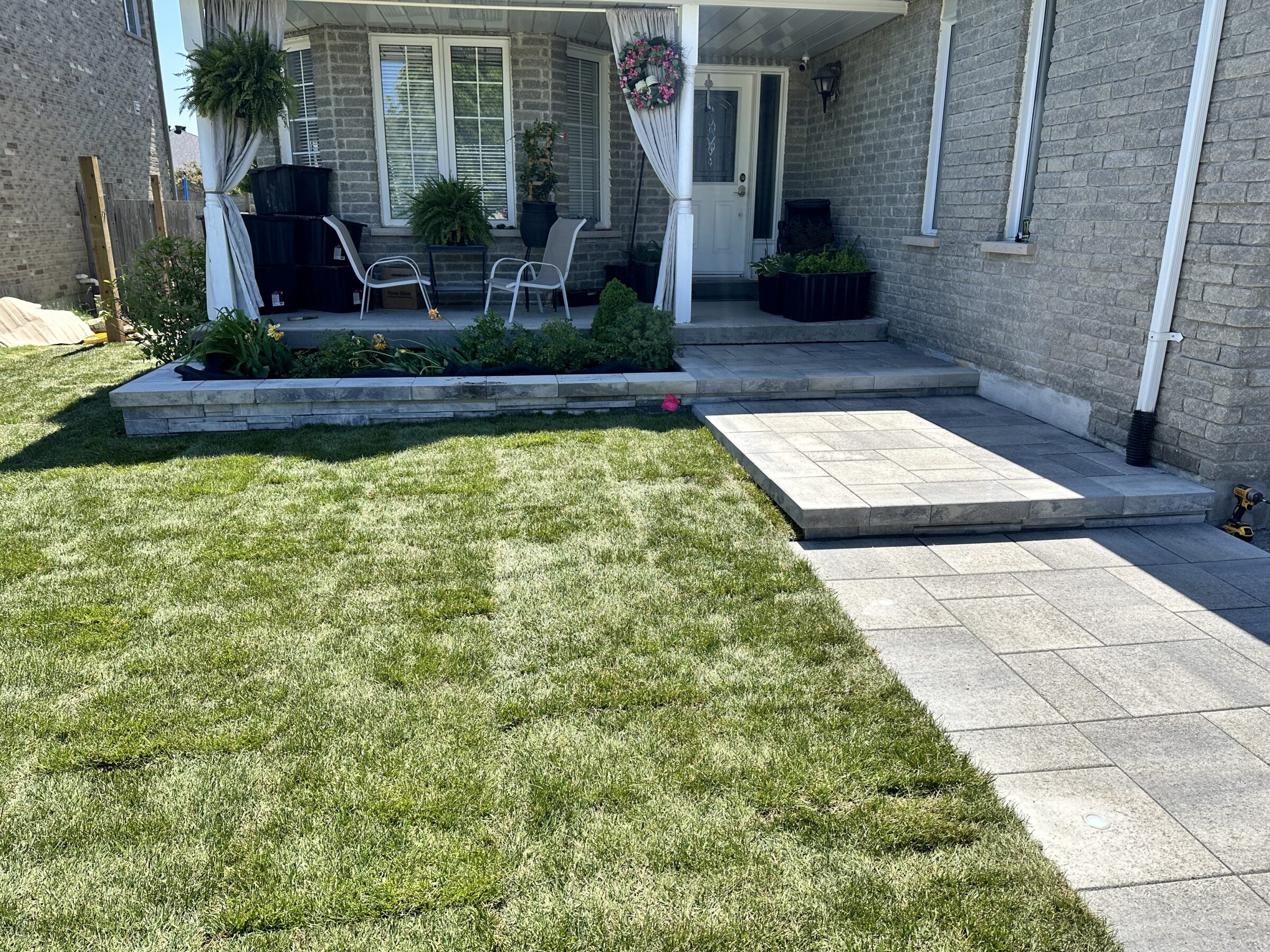 A neat garden with patio chairs, potted plants, and paved walkway outside a brick house, featuring a small porch area.