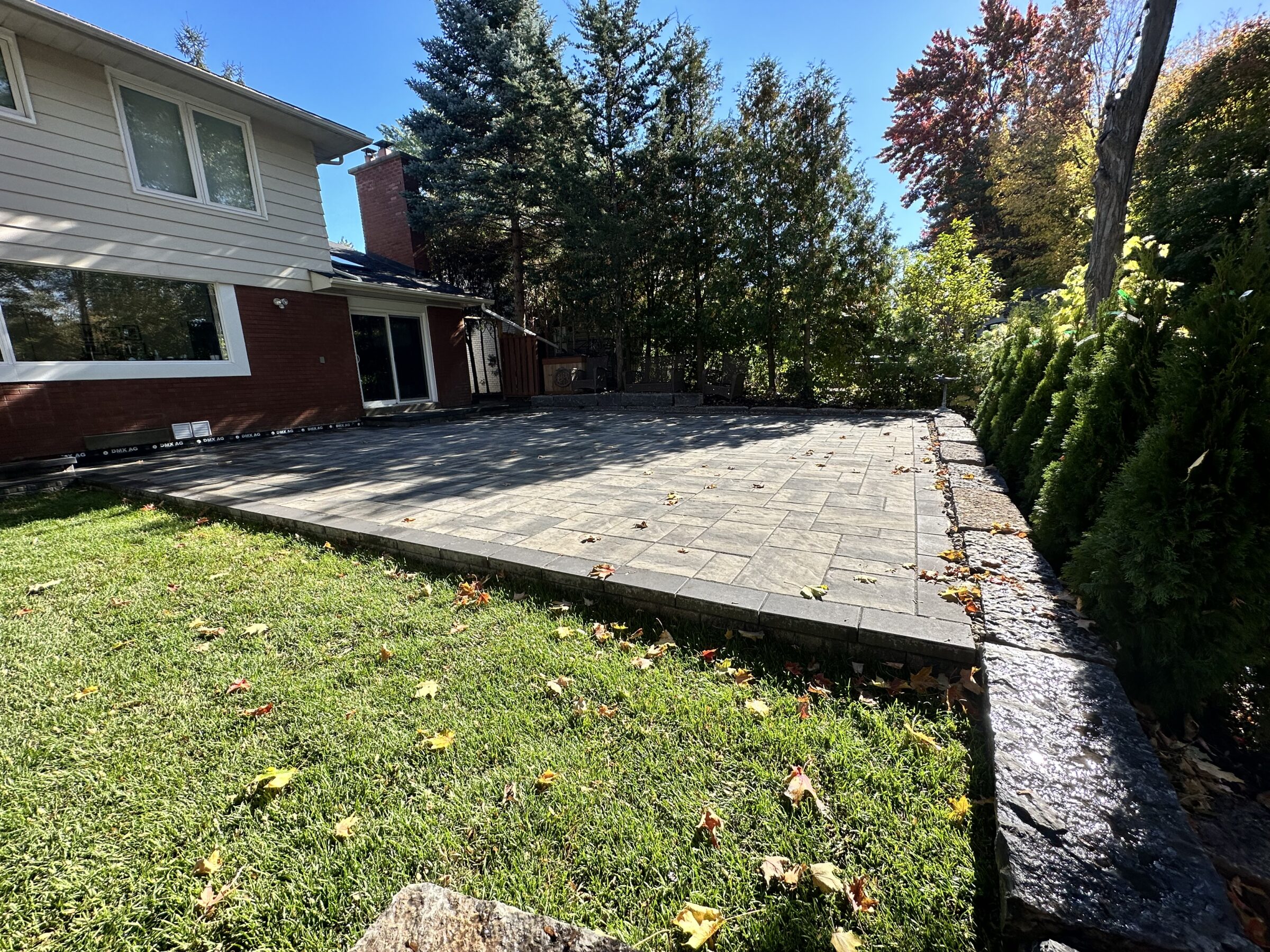 A large backyard features a paved patio, trimmed hedges, lush grass, and a view of a two-story house with large windows.