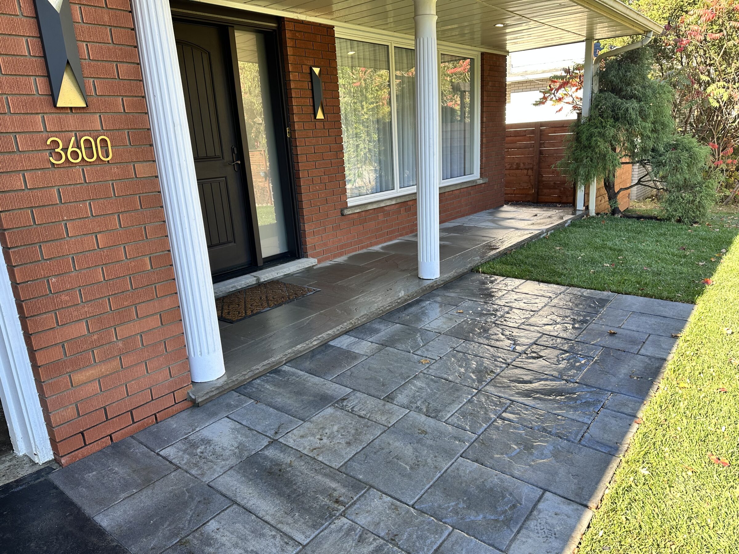 Brick house entrance with white columns, black door, and address number 3600. Stone path leads through a neatly landscaped, grassy lawn.