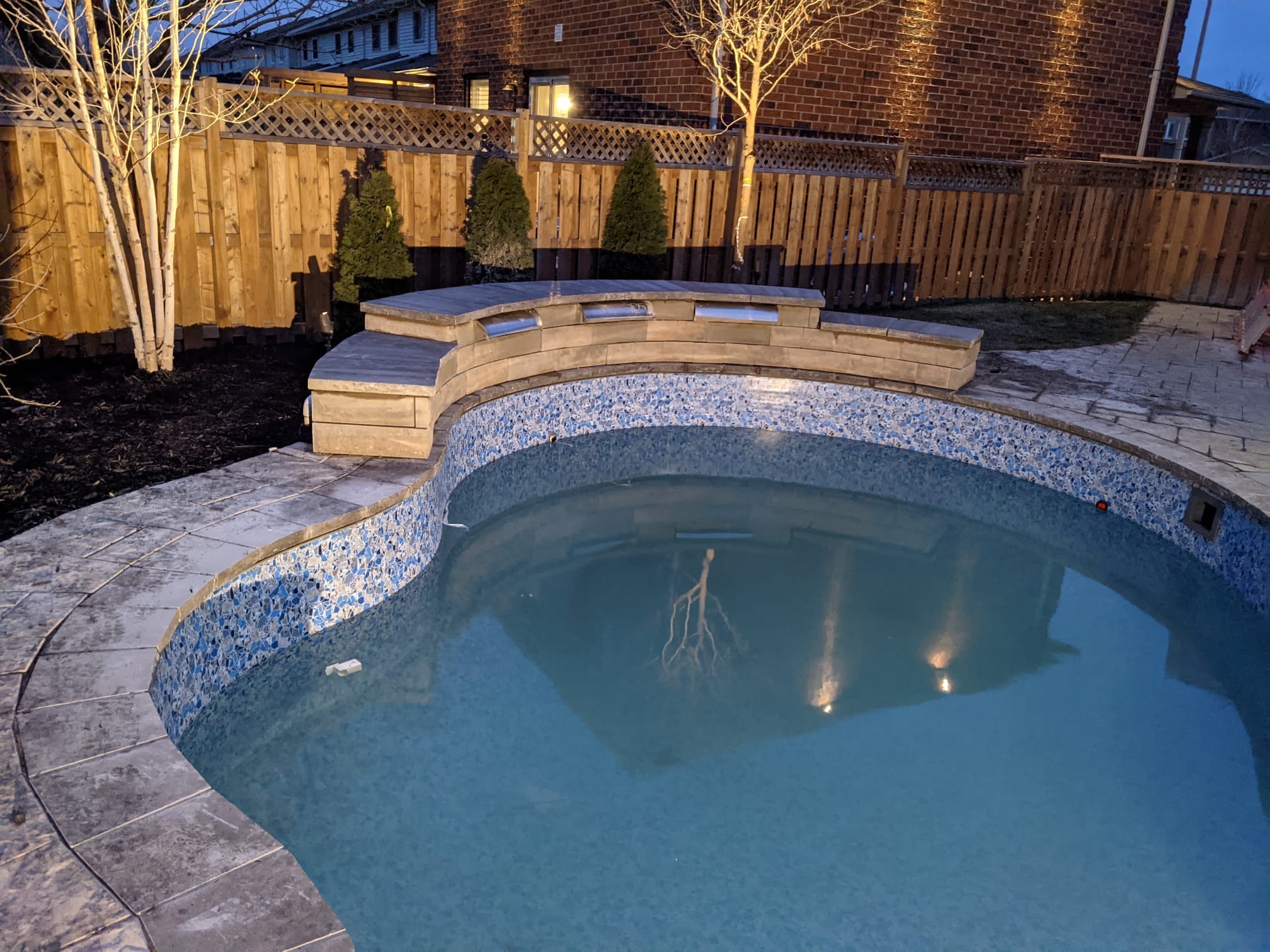 A backyard pool with mosaic tiles, surrounded by a wooden fence, trees, and decorative lights on a brick house exterior at dusk.