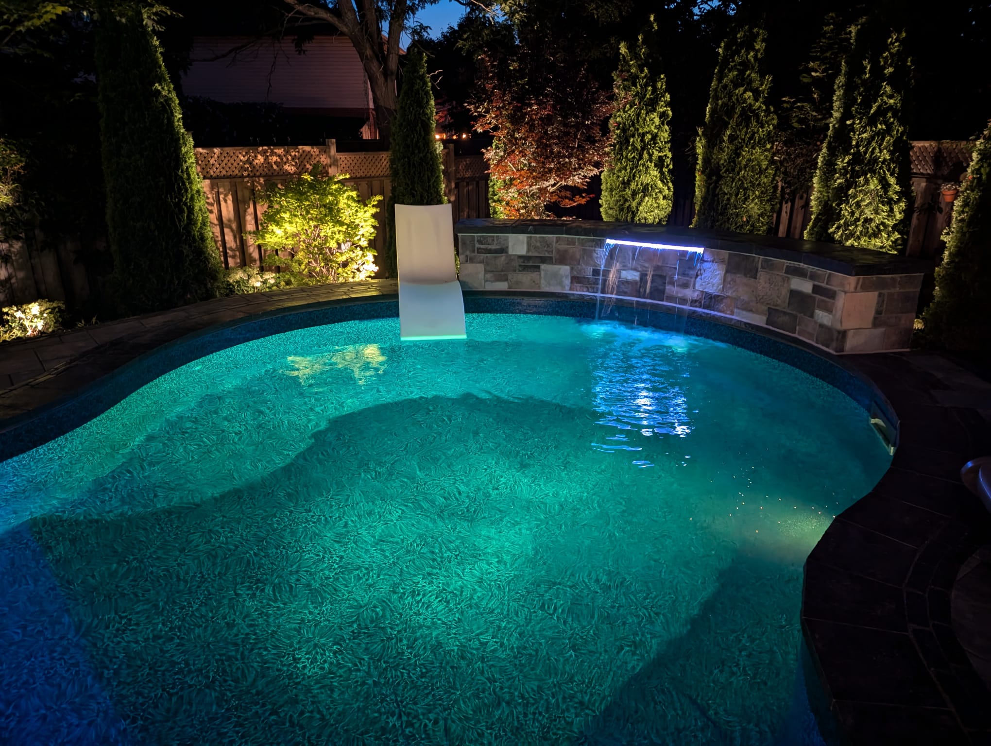 A beautifully lit backyard pool at night, surrounded by tall trees and illuminated water features. Tranquil and inviting ambiance.